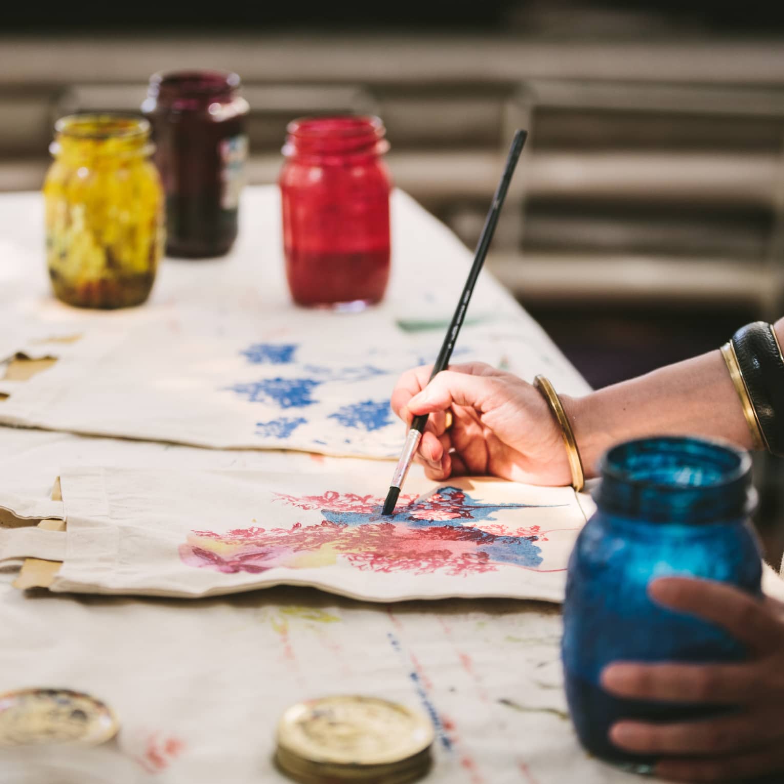 An artist using mason jars filled with red, blue, yellow and black paint to paint images of flowers on canvas tote bags.