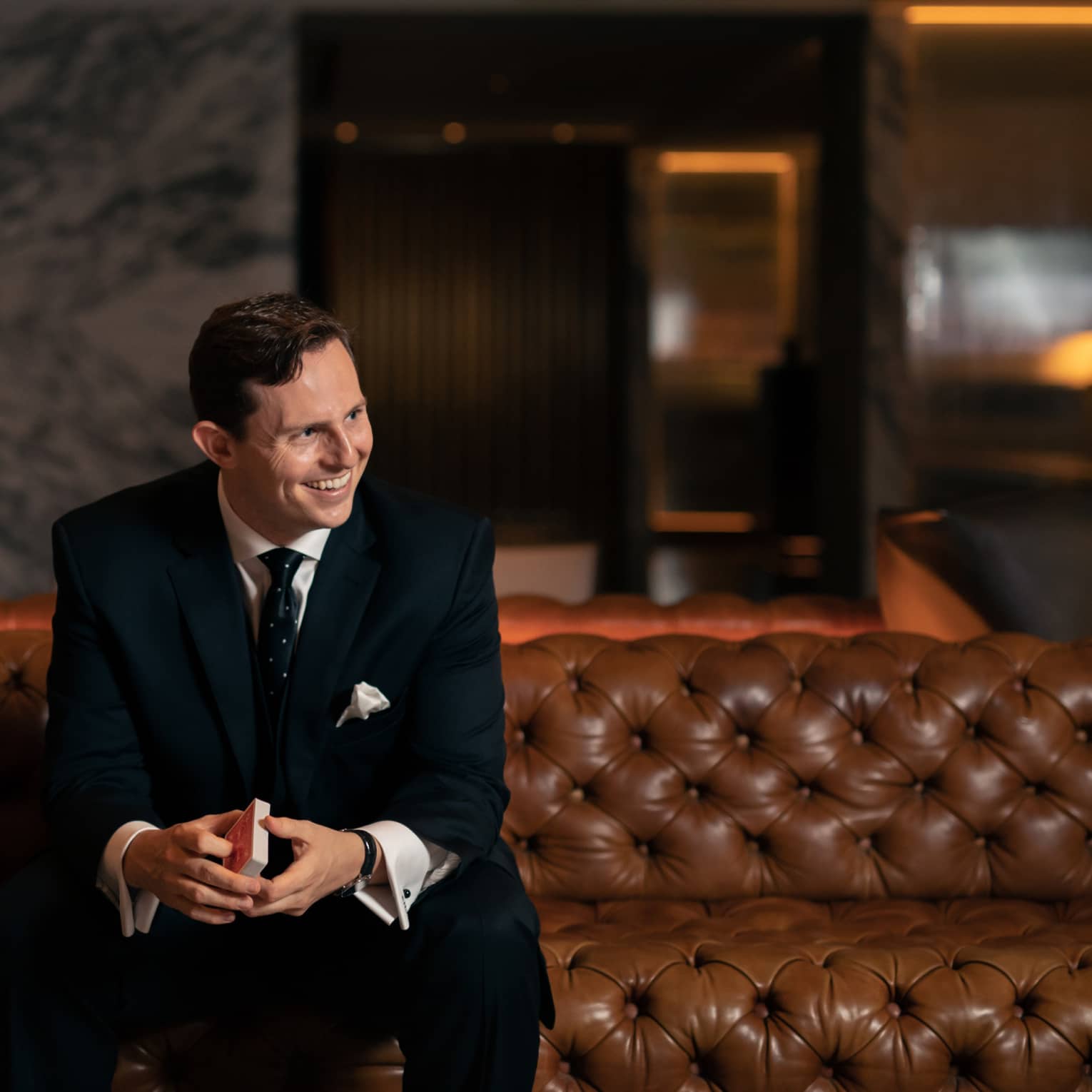 A man in a black suit sitting on a leather sofa holding playing cards.