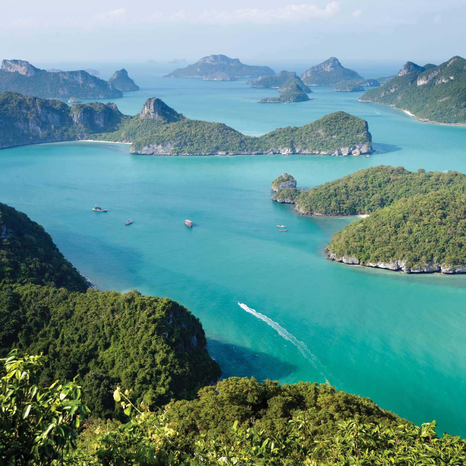 Aerial view of a dozen lush, forested, hilly islands of different sizes surrounded by calm turquoise water dotted with boats.