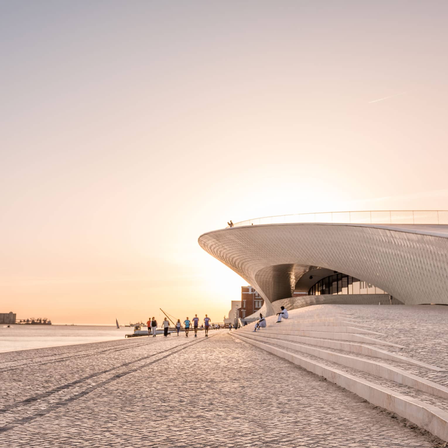 Modern waterfront building with sweeping architectural curves, people walking along a cobblestone promenade at sunset.