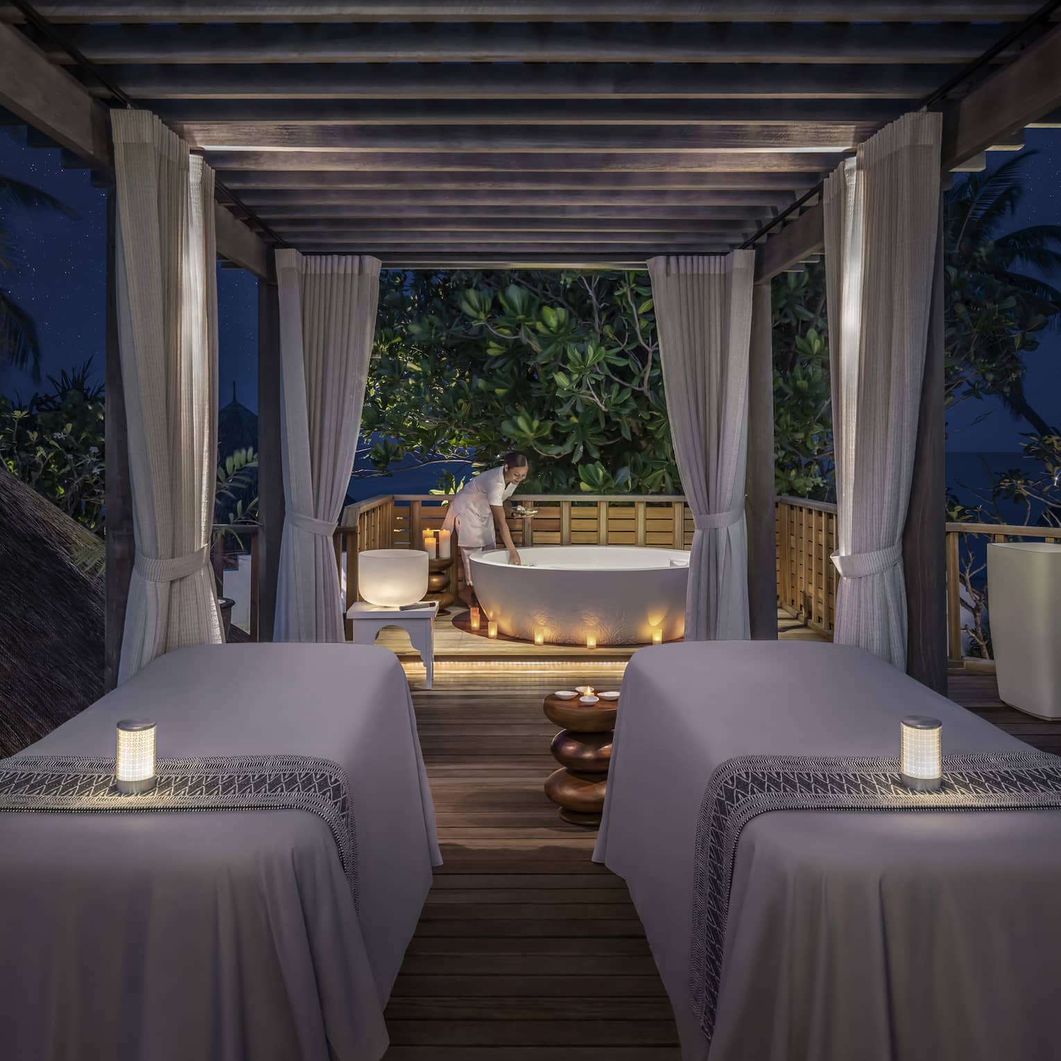 Outdoor night spa setting with two massage tables, soft candlelight, and a staff member preparing a bathtub under a canopy surrounded by tropical greenery