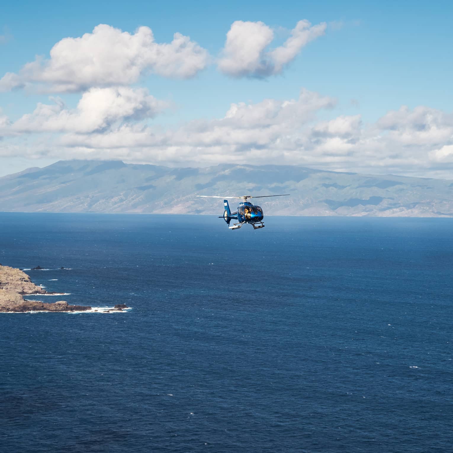 A helicopter flies over open ocean with a mountainous island and blue sky dotted with clouds in the background