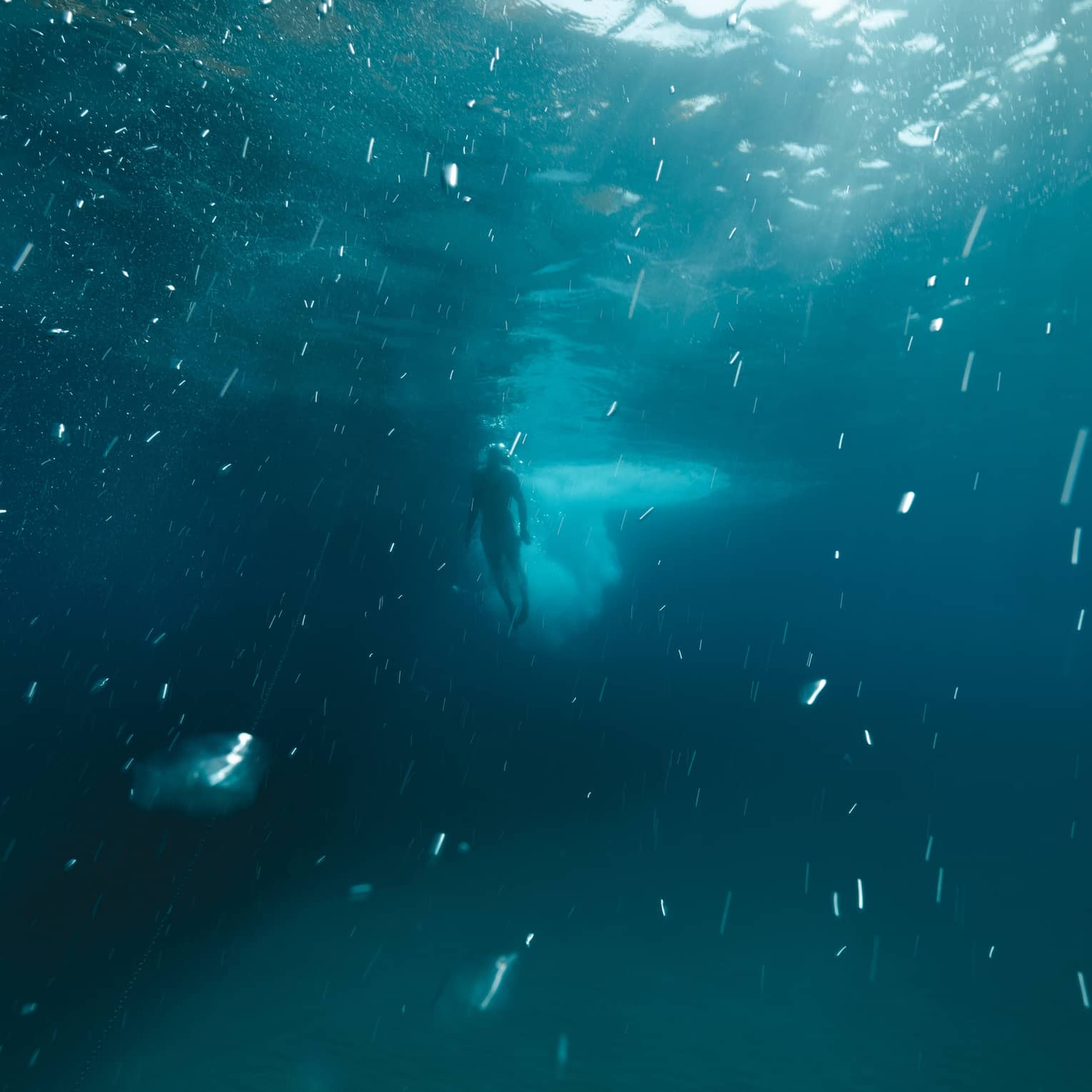 Deep underwater long view of a diver swimming toward the surface as sunlight pierces through the dark blue water.