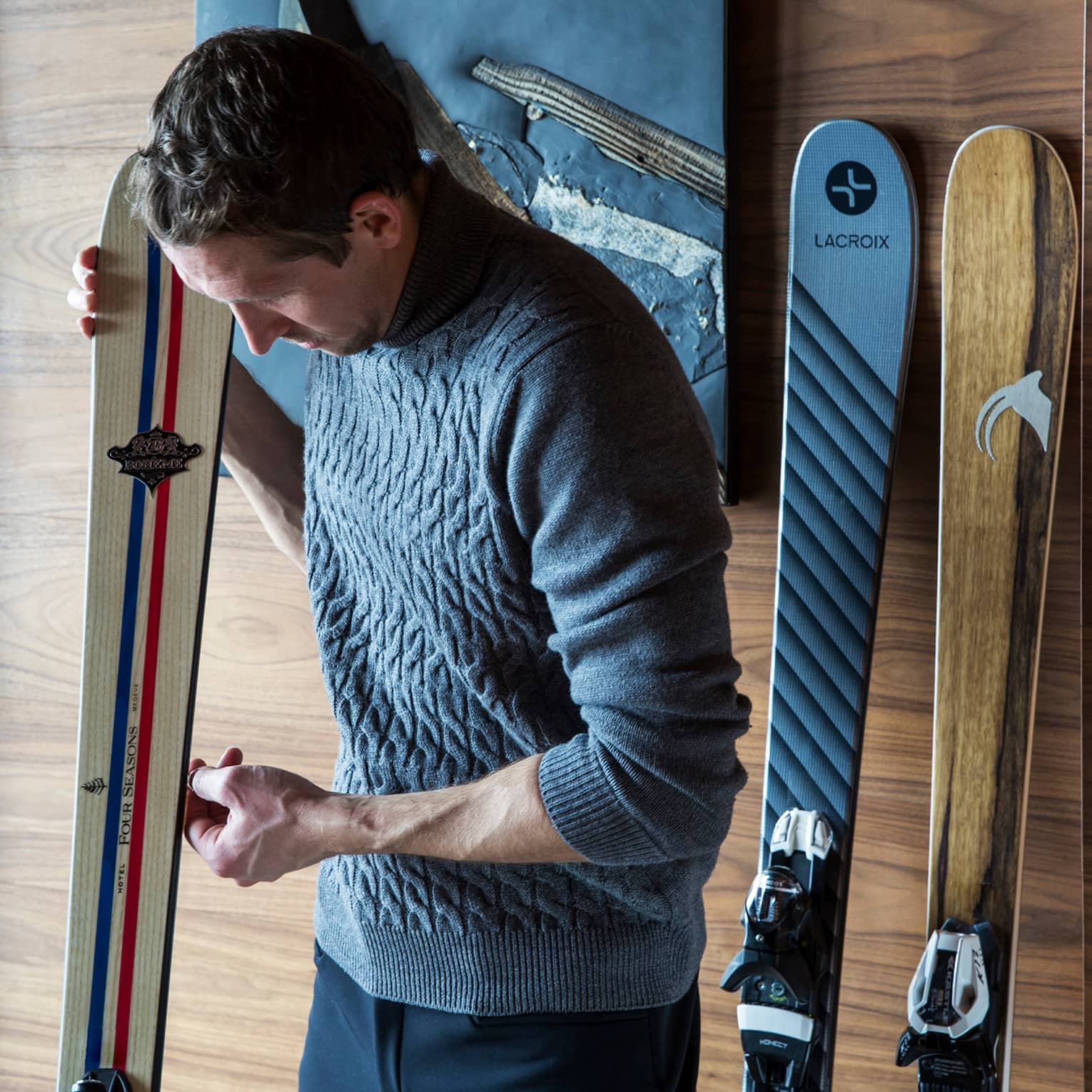 Man wearing knit sweater leans over ski against wall, more skis on wall