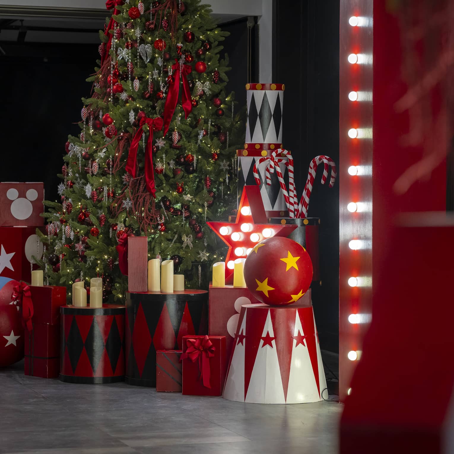 In a dark room, a tall Christmas tree is surrounded by candles, large candy canes, and wooden boxes, cylinders and balls.