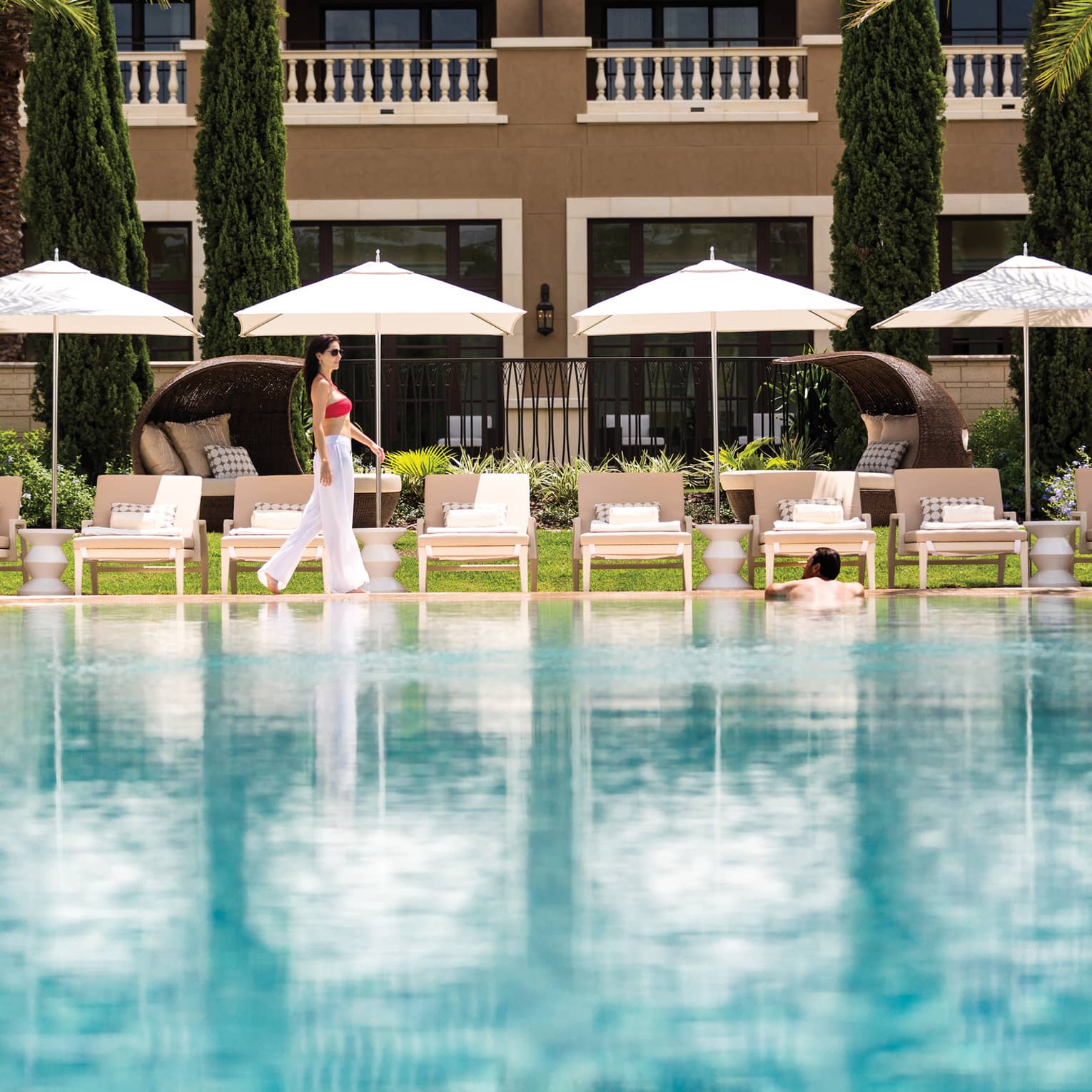 Woman walks by row of white patio chairs, umbrellas on outdoor swimming pool deck
