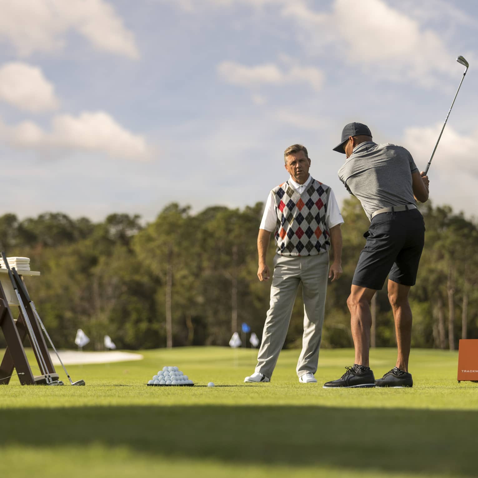 Against clear sky, one golfer looks on as another prepares to swing, a tray of balls and a stand of clubs nearby.