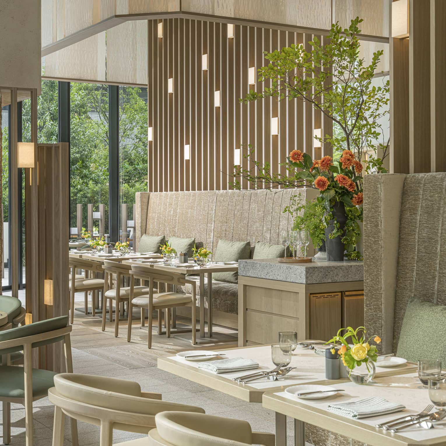 Airy earth-toned bistro with green cushions on tall banquettes, vases of flowers on tables and windows showing lush foliage.