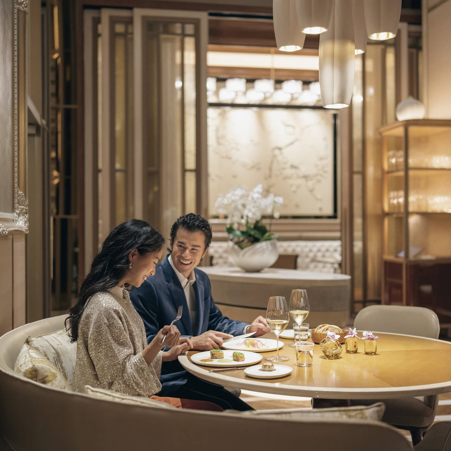 A smiling couple savours lunch with wine in a circular booth at a fine dining restaurant with warm pendant lighting.