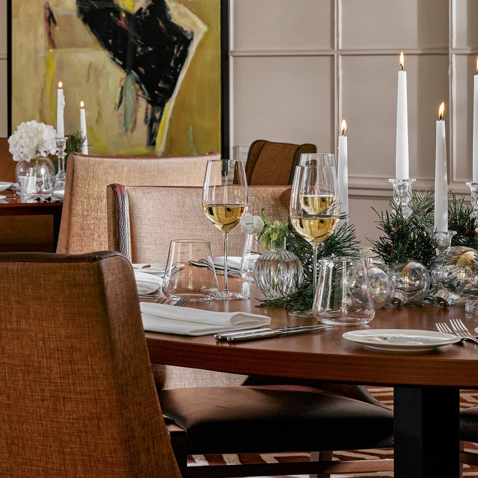Elegantly set holiday table with greenery, lit white taper candles, glasses of white wine and white porcelain dishes