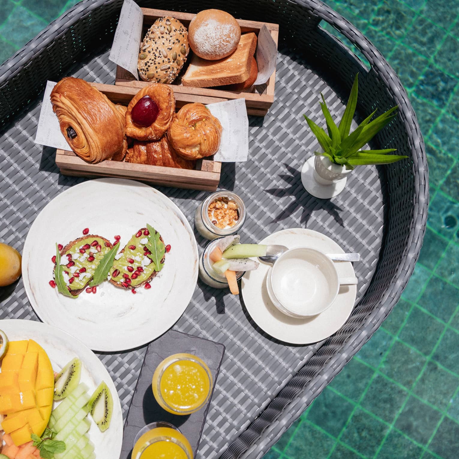Close-up view from above of a wicker tray filled with fruit, pastries, juice and toast floating in a crystal-clear pool.