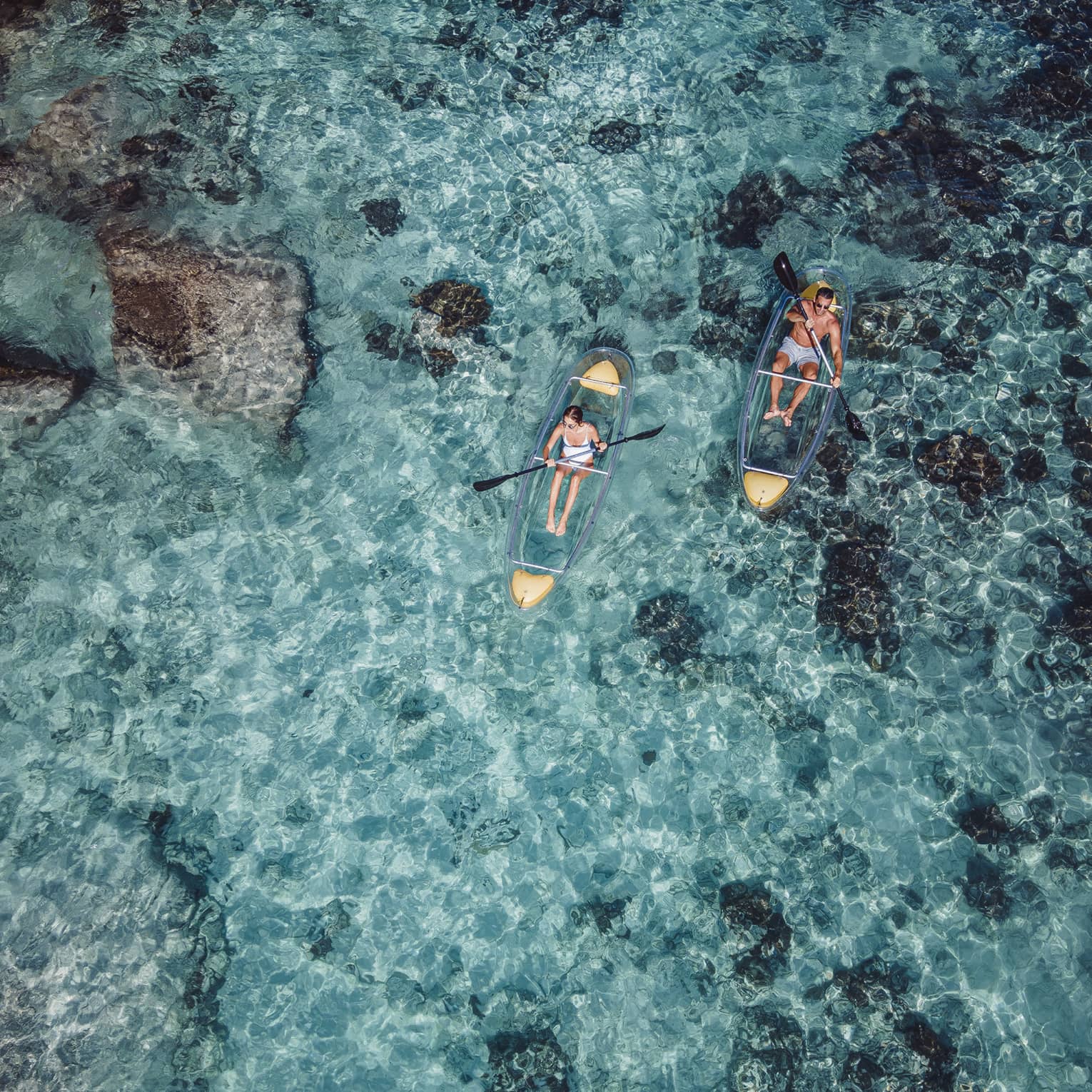 A view from above of kayakers paddling in transparent kayaks near boulders jetting out of crystal-clear open water.