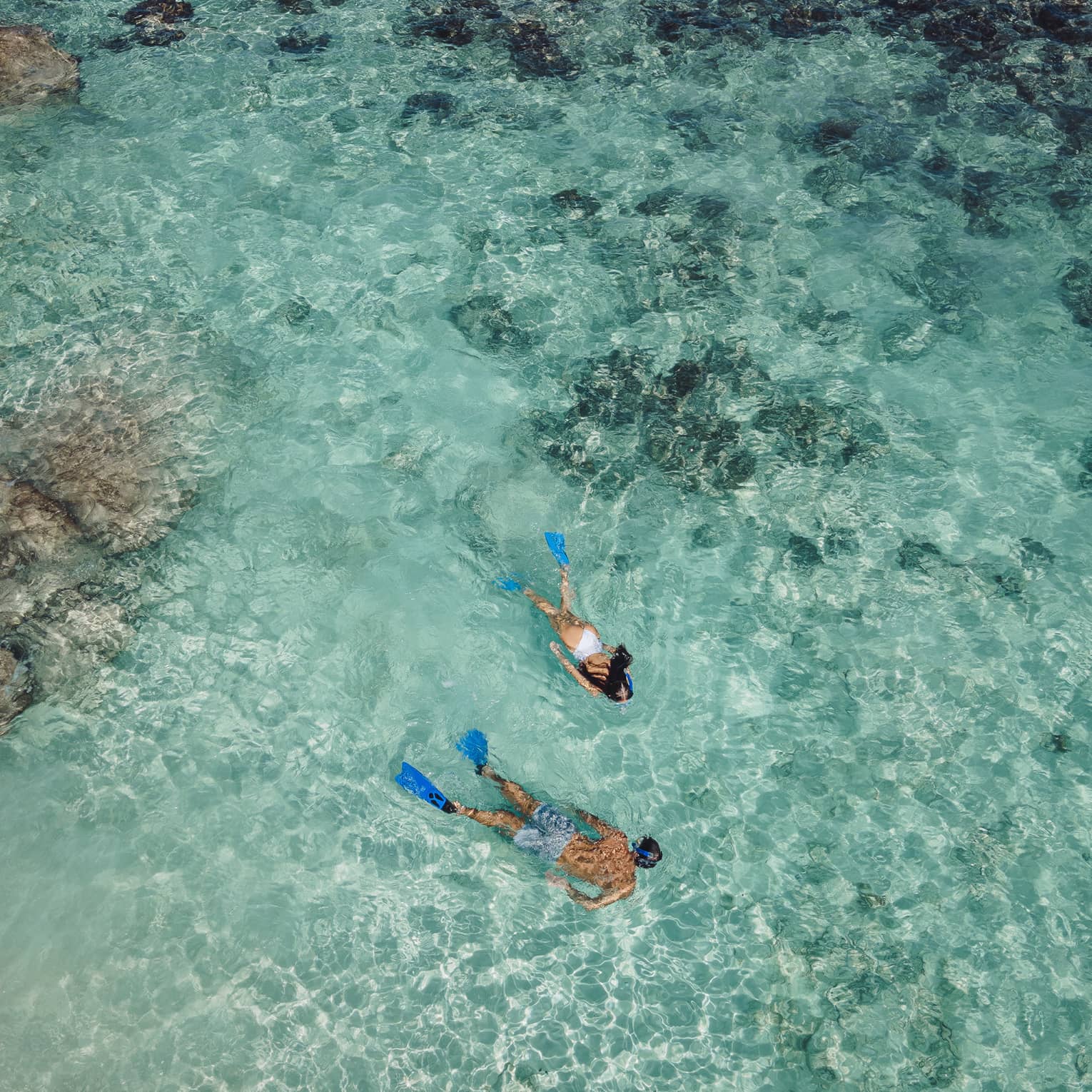 Aerial view of two snorkellers wearing blue flippers in an expanse of shallow, turquoise waters rippling over the reef.