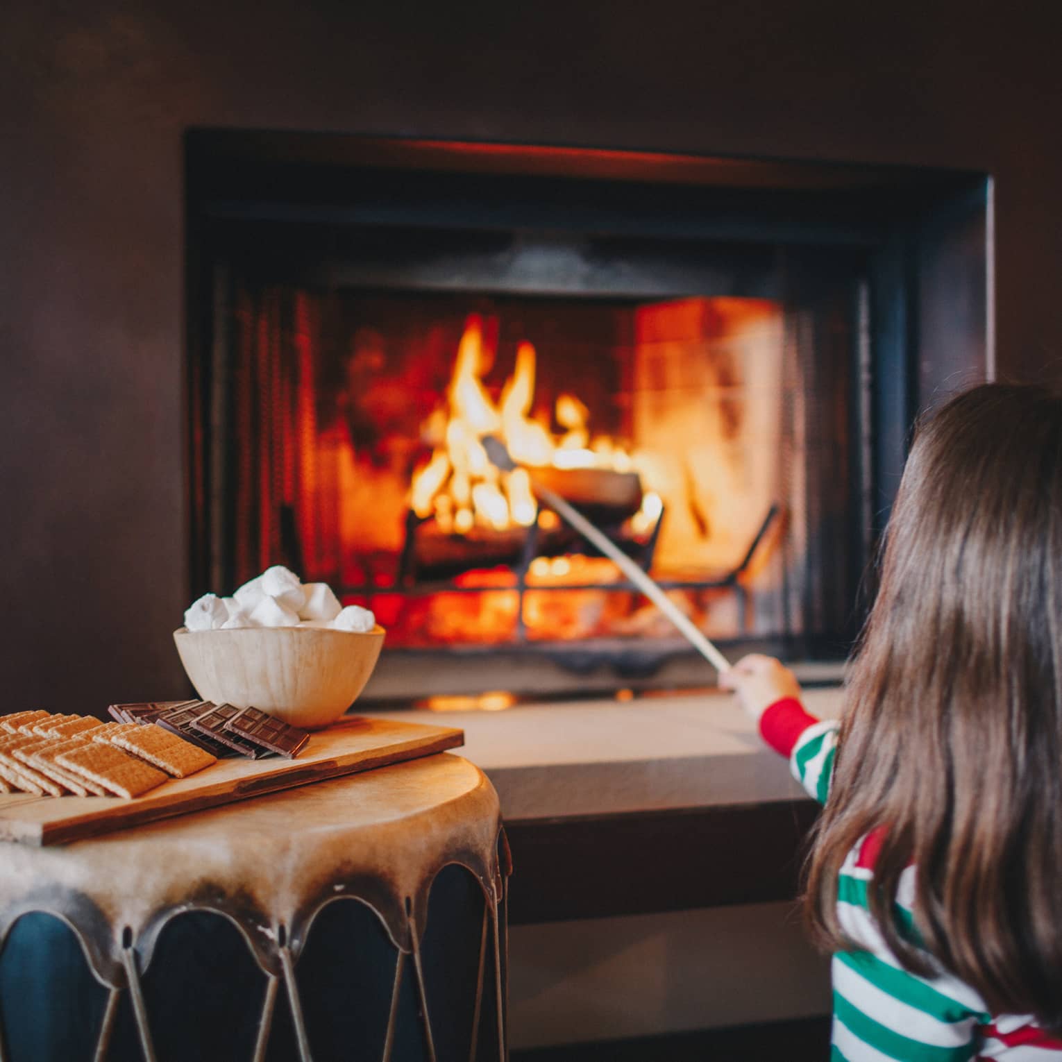 A little girl in red and green pajamas roasts marshmallows in a fireplace