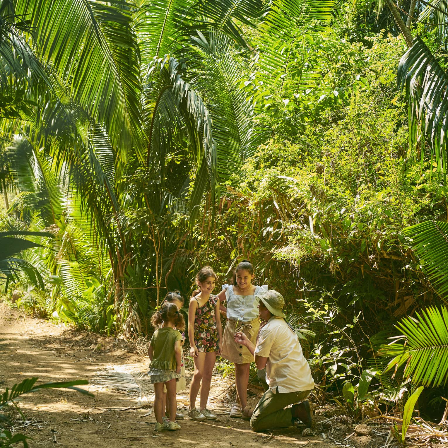 A guide wearing a sun hat kneels before a group of smiling children on a sun-dappled dirt trail amidst lush green foliage.