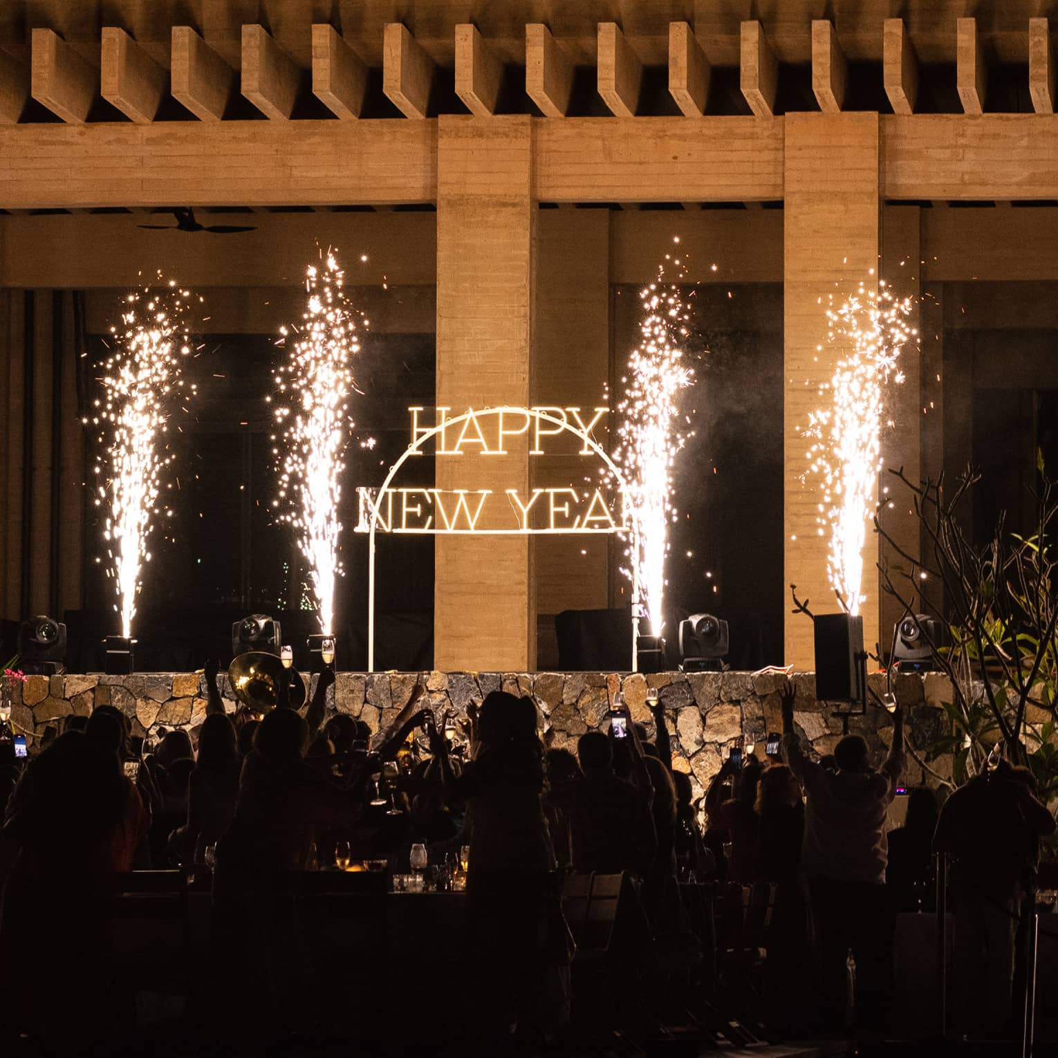 A glowing Happy New Year sign with sparklers beside it, and a crowd cheering.