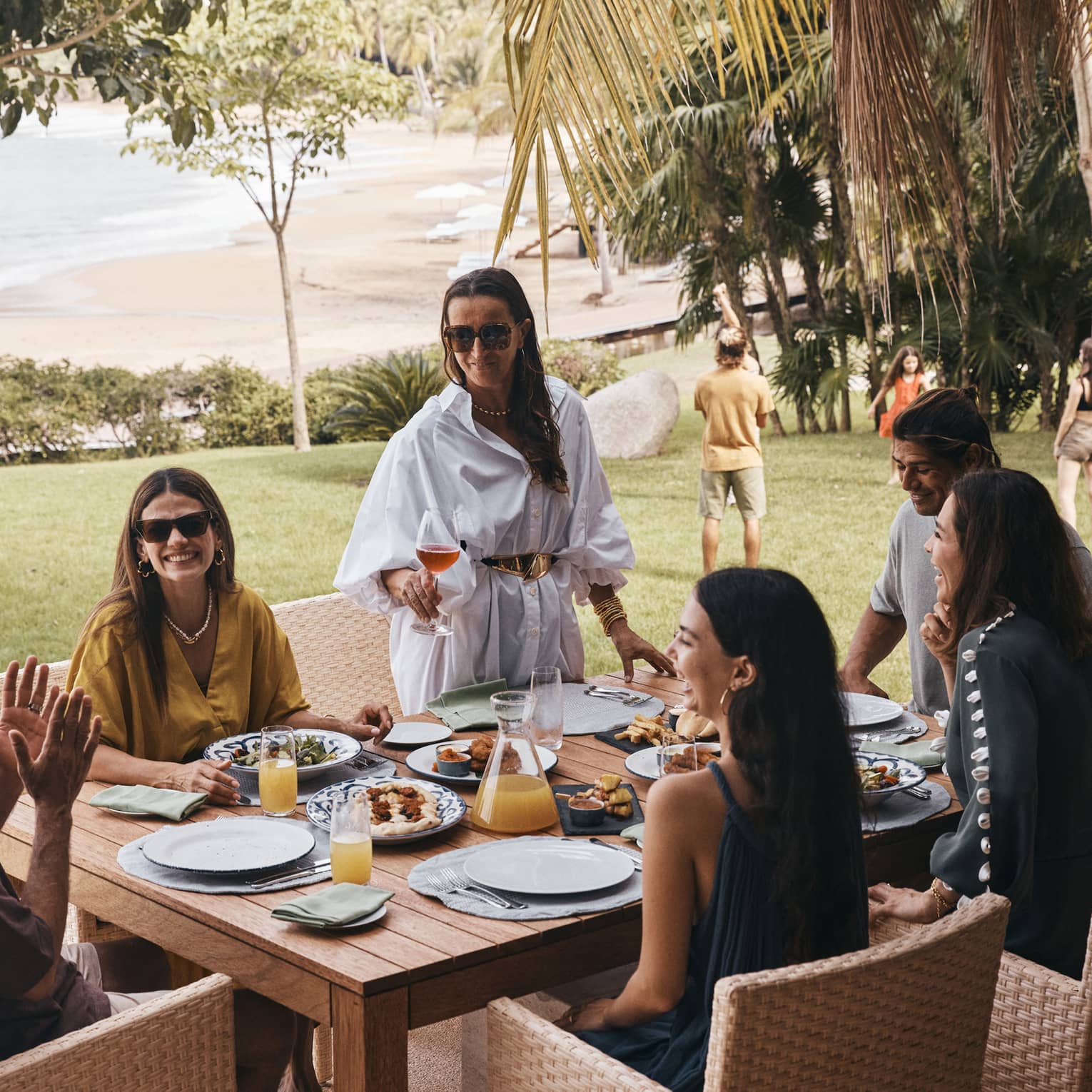 People seated around a square wood dining table in an outdoor area surrounded by trees and palm trees, with a lawn, beach and the shoreline in the background.