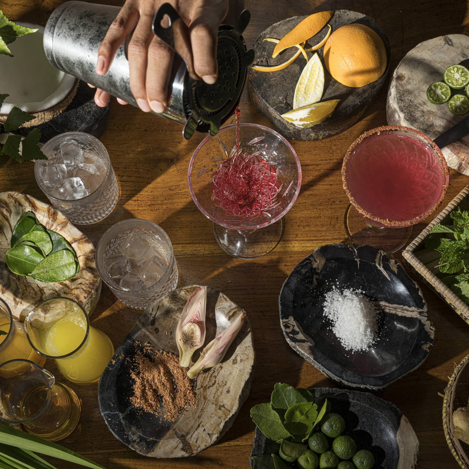 Aerial view of bartender's hand pouring ruby-coloured cocktail intro stemmed glass, local ingredients on wooden bartop surrounding it