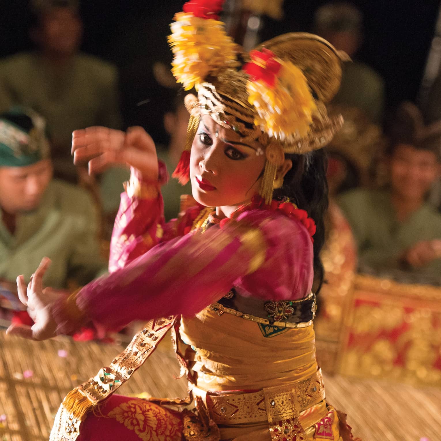 Close-up of a Balinese dancer in a golden headdress and bright clothing; in the background musicians play the gamelan.