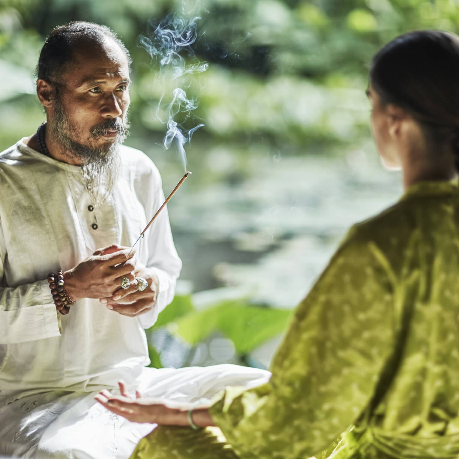 Set against a backdrop of greenery, Djik Dewa holds a stick of burning incense before a meditative cross-legged guest.