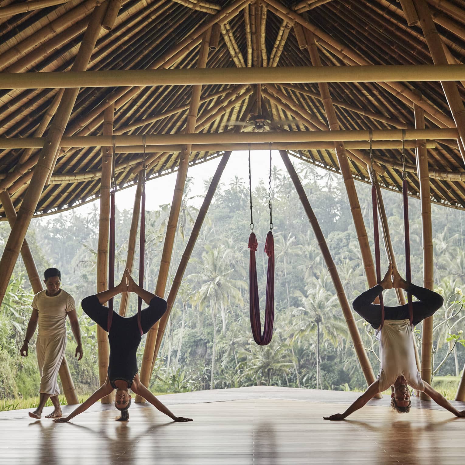 Woman and man hang upside-down on yoga hammocks under wood pavilion as instructor looks on