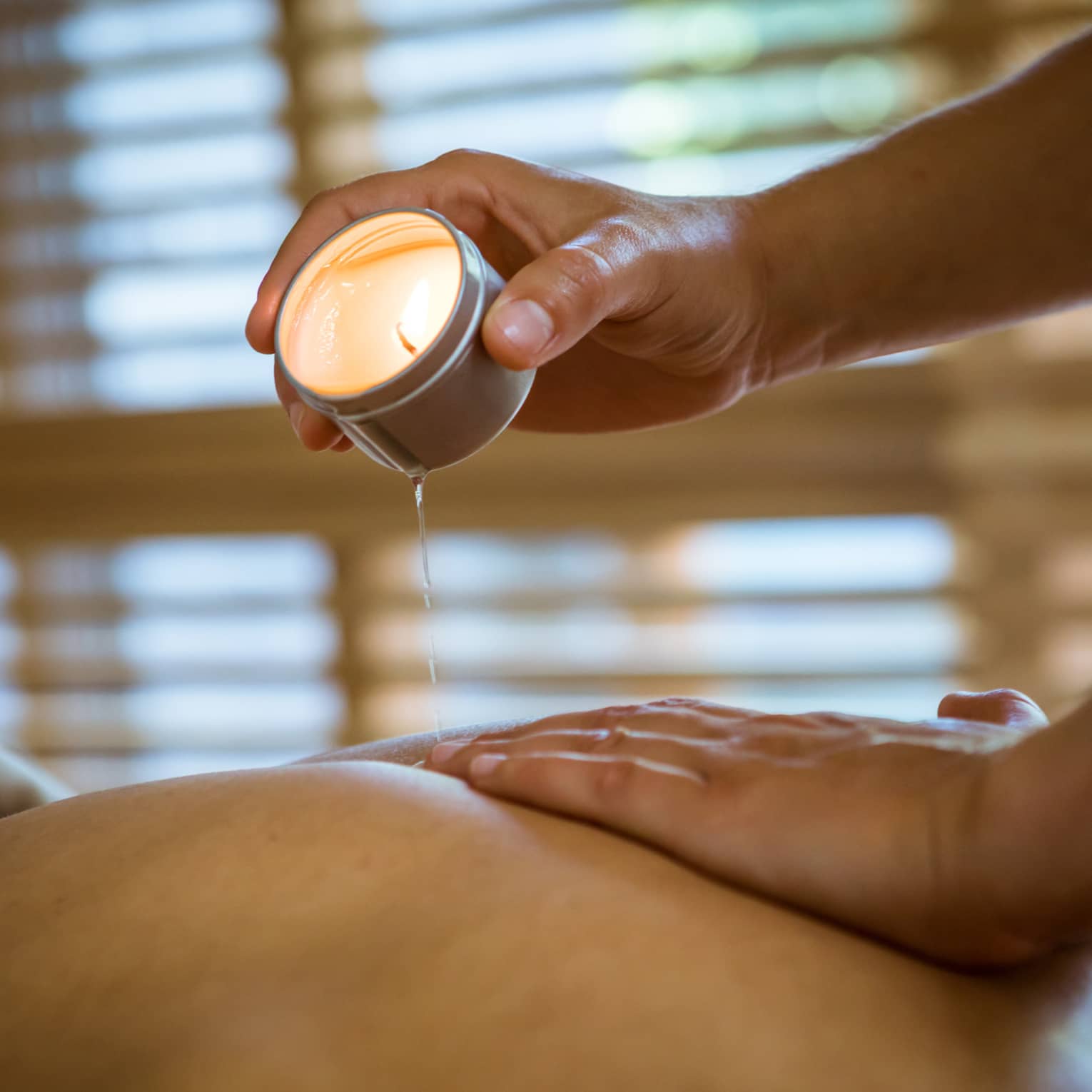 Massage therapist pours hot wax from tealight candle over person's bare back in spa