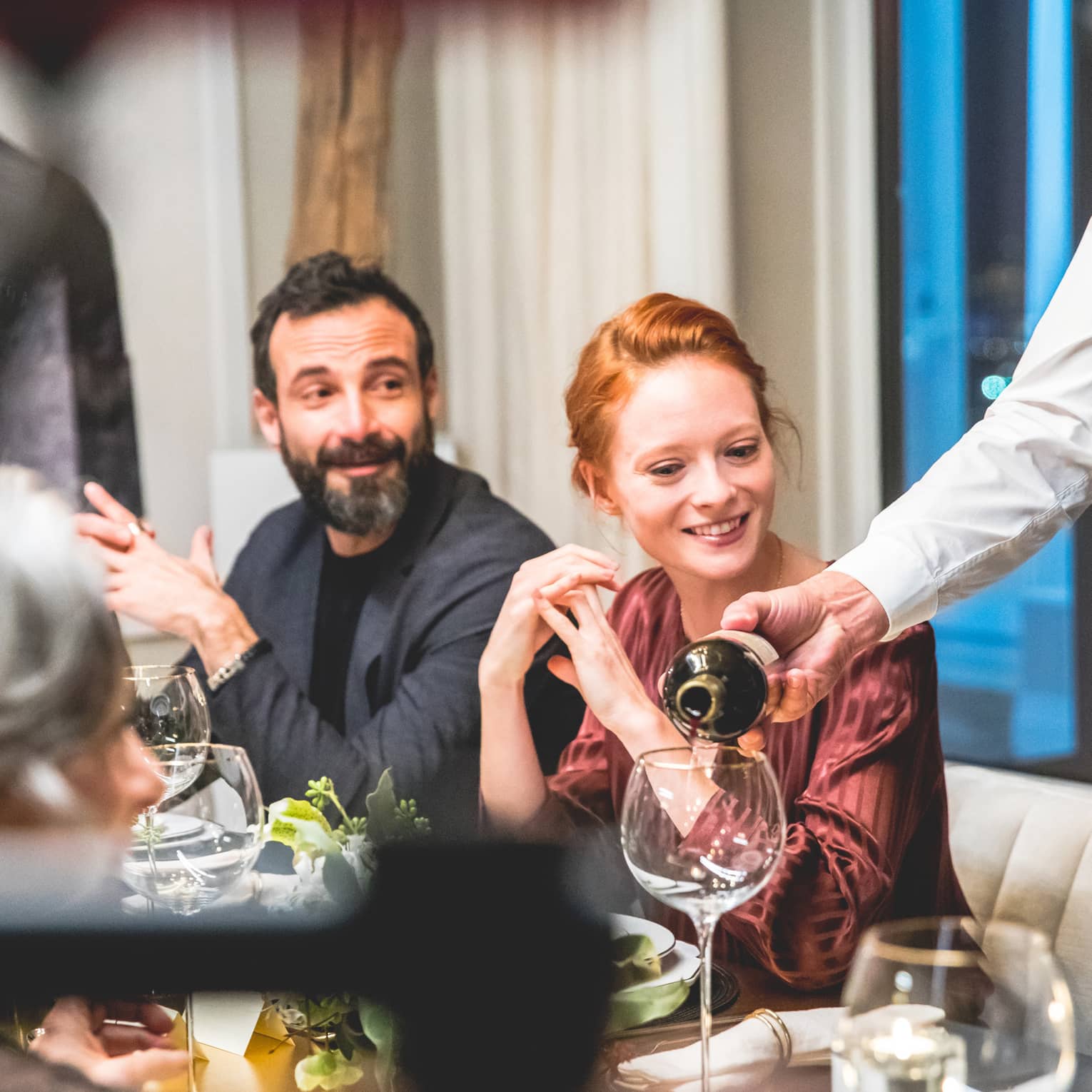 A man and woman being served a red wine at a well lit restaurant.