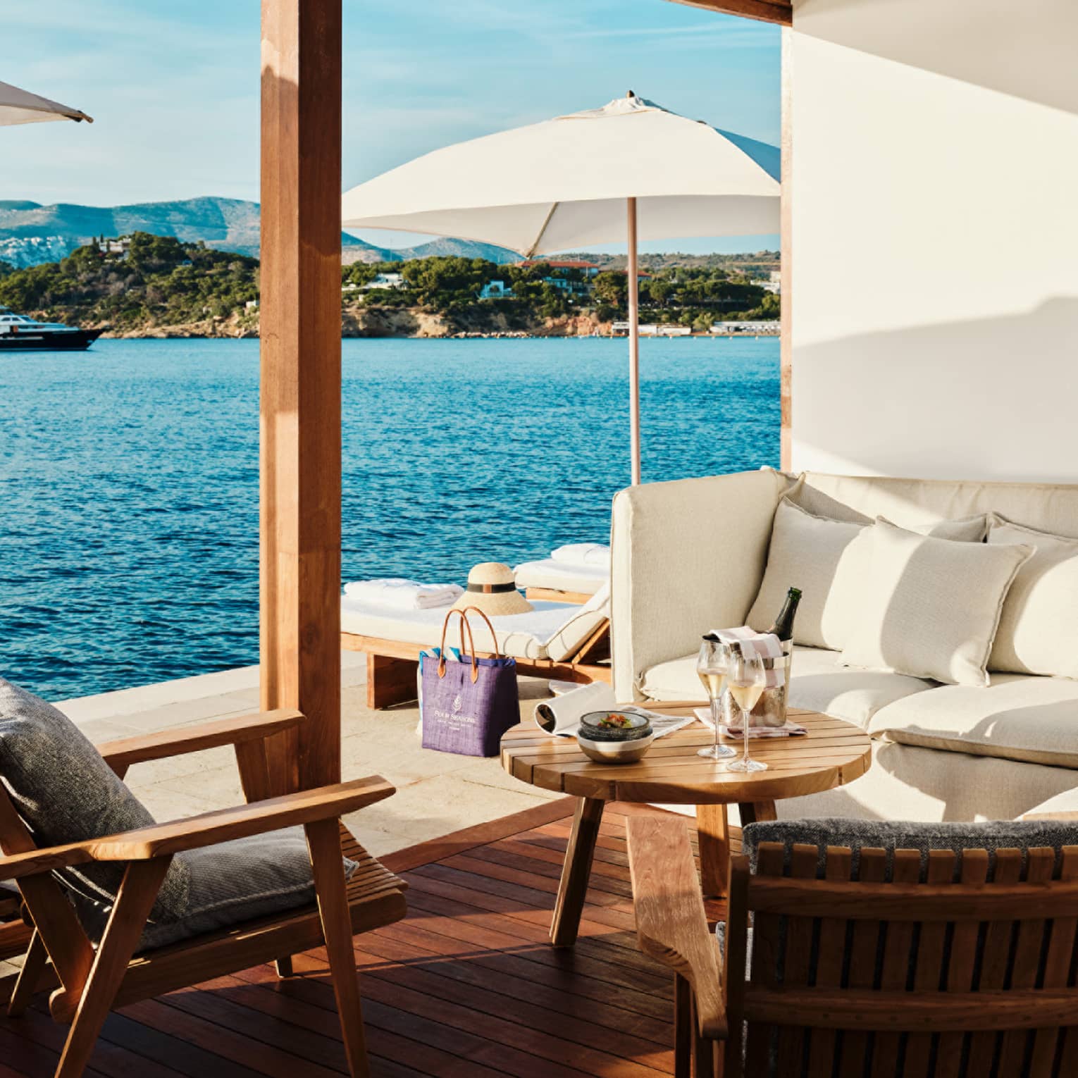 Cabana interior with white banquette-style couch, round teak table and two teak chairs open to platform with white umbrellas and blue sea views