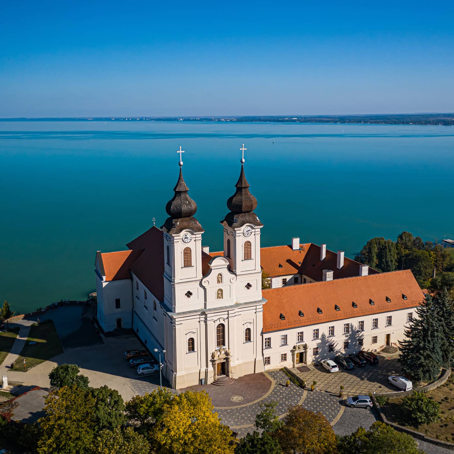 A Benedictine monastery, with white walls and a red tiled roof, sitting beside a large blue lake and surrounded by trees.