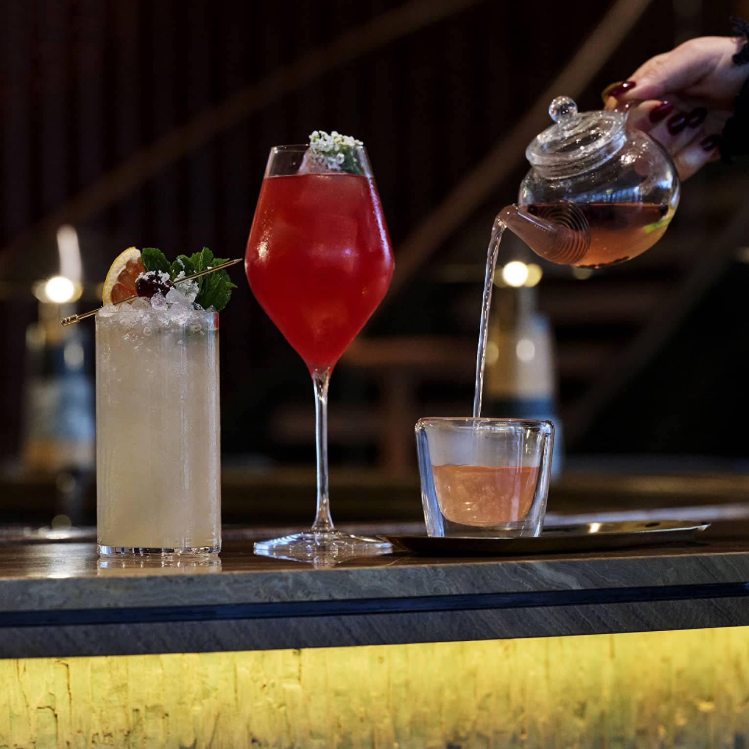 Two cocktails shown on a bar next to another beverage being poured into a glass from a small teapot