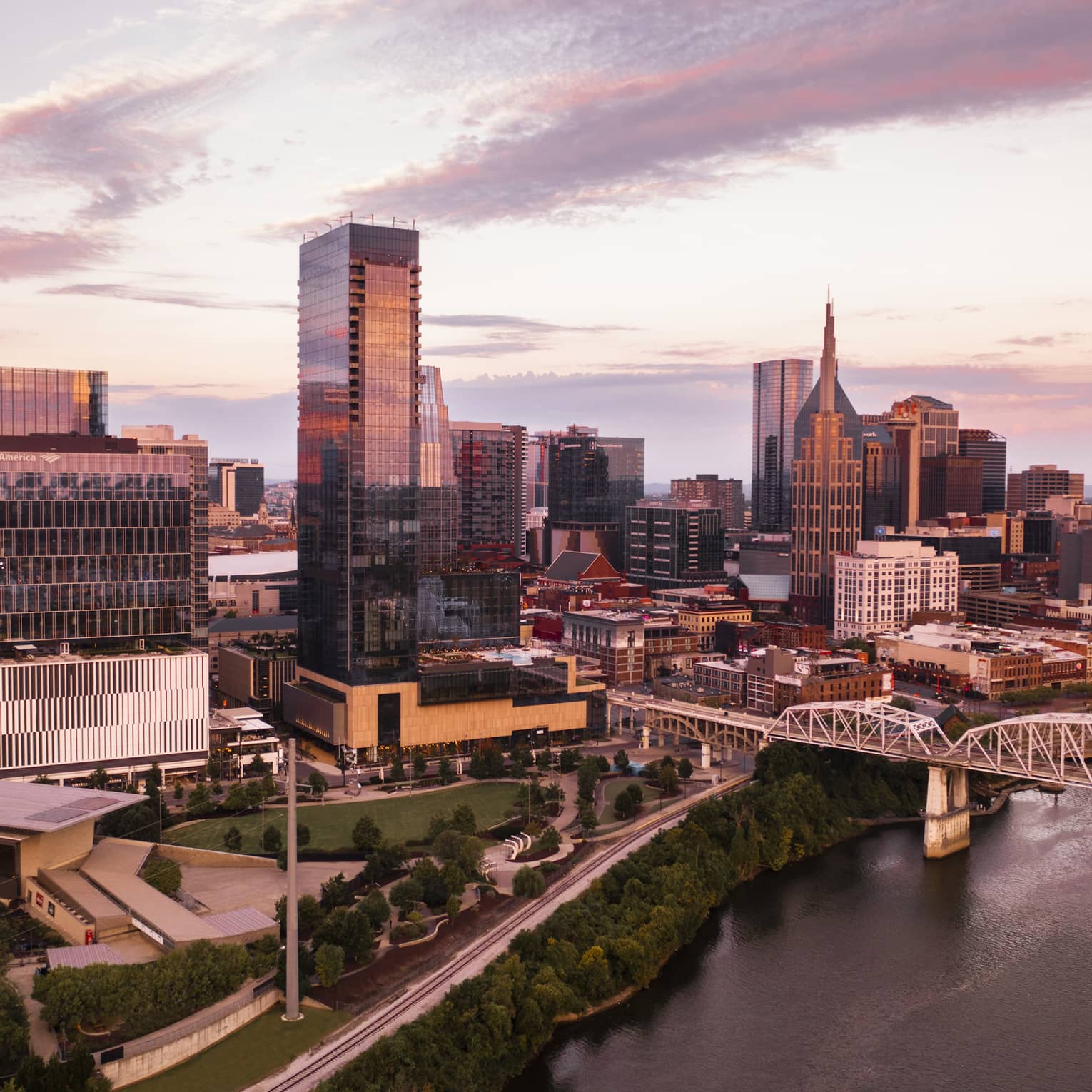 Aerial view of downtown Nashville, Tennessee at sunset, featuring modern skyscrapers, a river, a bridge, and a park with greenery in the foreground.