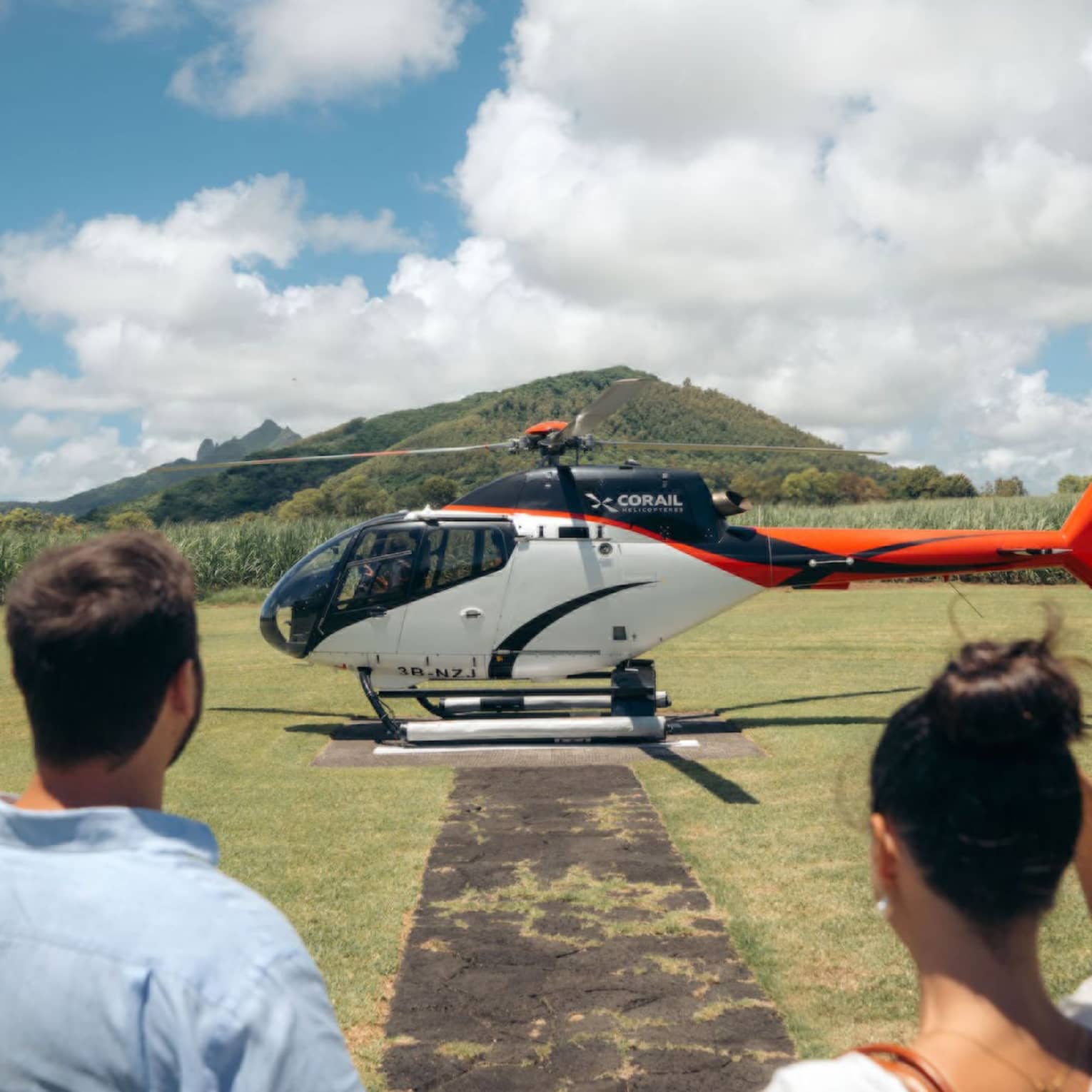 A couple is looking at a helicopter sitting on a helicopter pad in a grass clearing with tall vegetation encircling it.