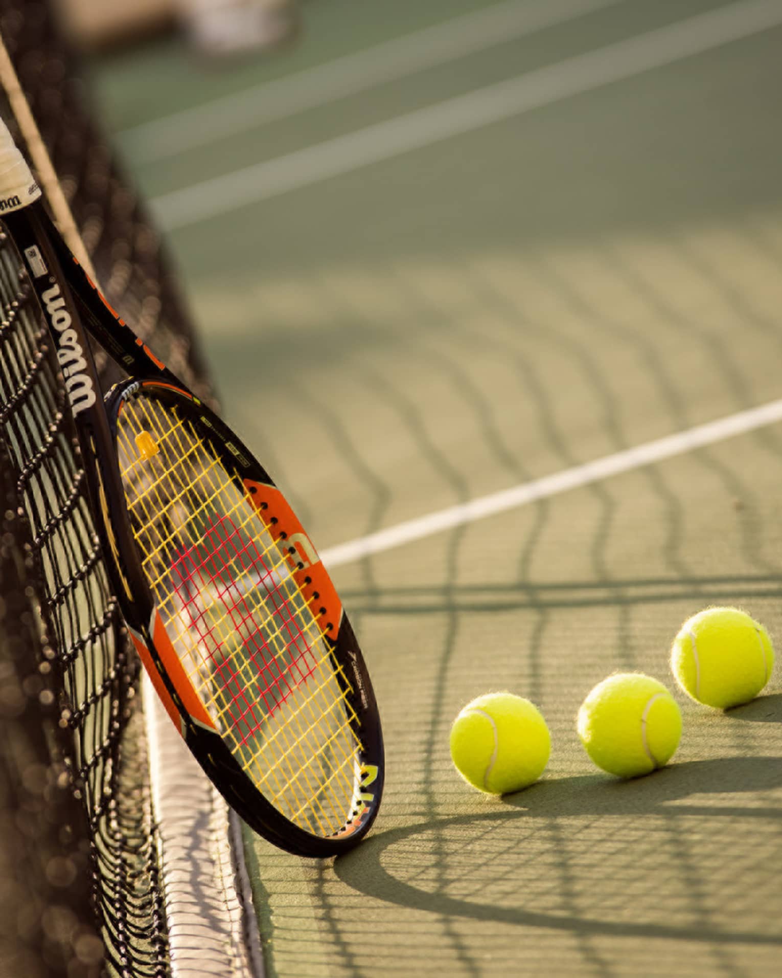 Red and black tennis racket leans against net by green tennis balls on court