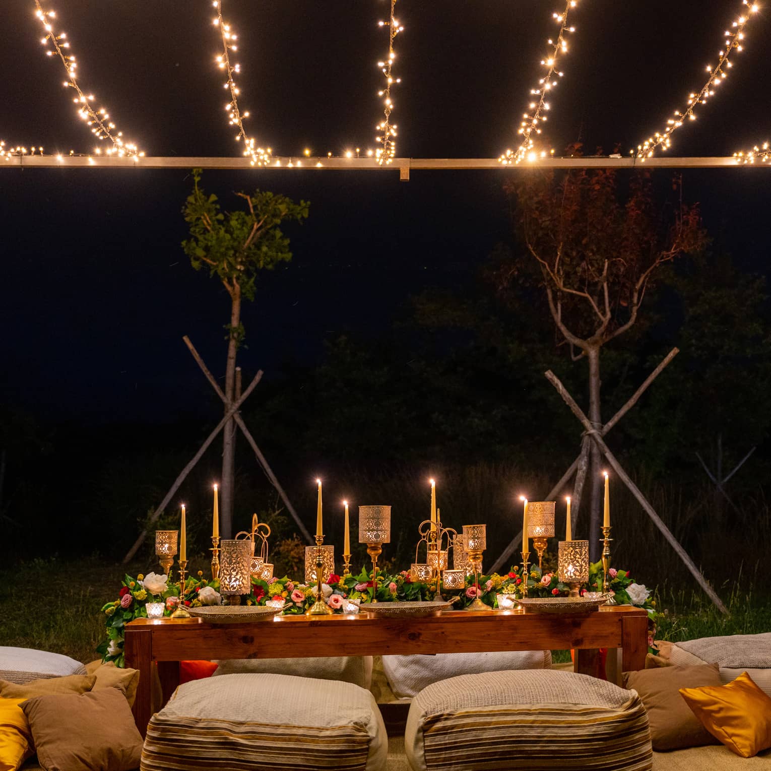 Low table with lit taper candles and gold lanterns, lush floral runner, surrounded by large cushions, under string lights.