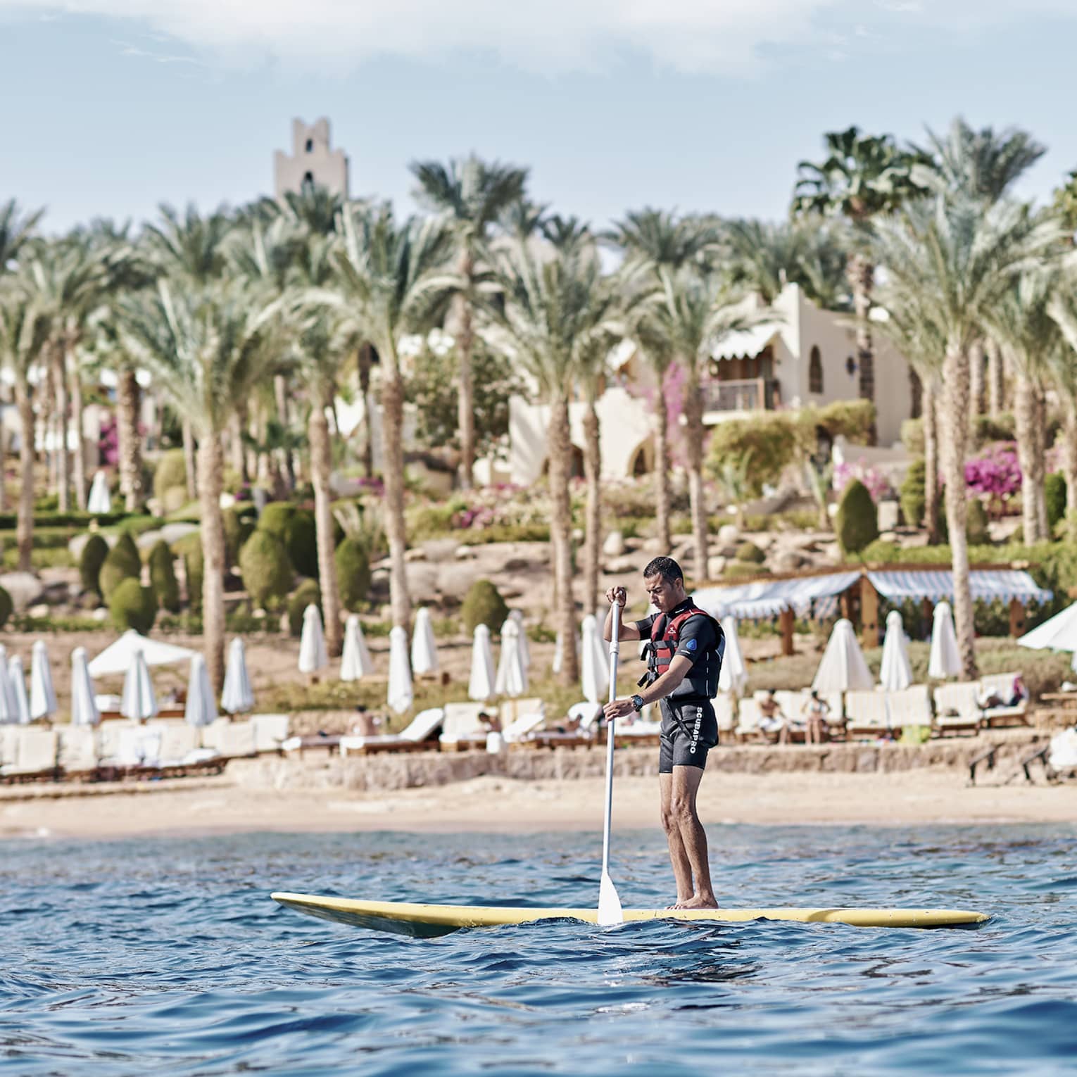 A paddler stands on a board in the water in front of a beach lined with lounge chairs and palm trees and a villa beyond.
