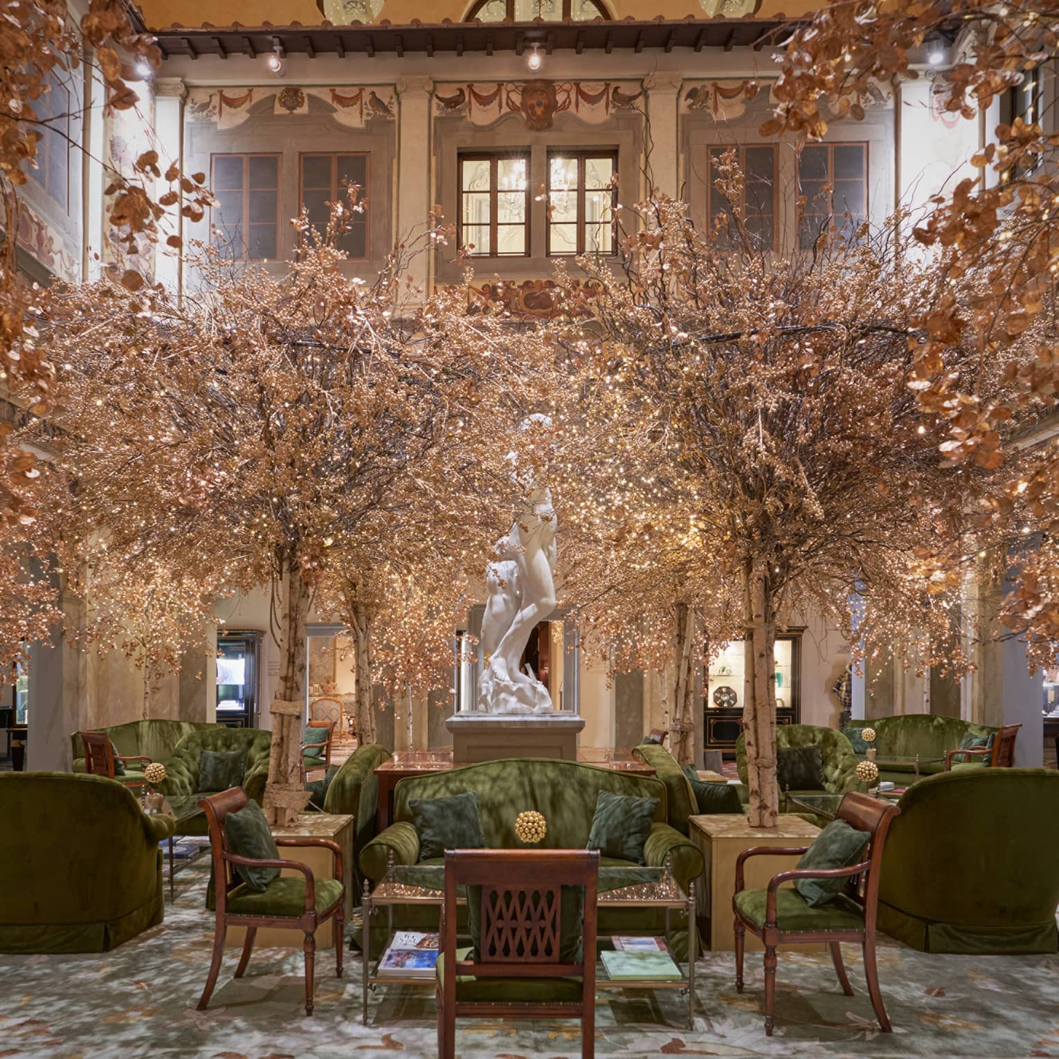 A large two-storey lobby is decorated with green velvet couches and chairs, golden-leafed trees, and a white marble statue.