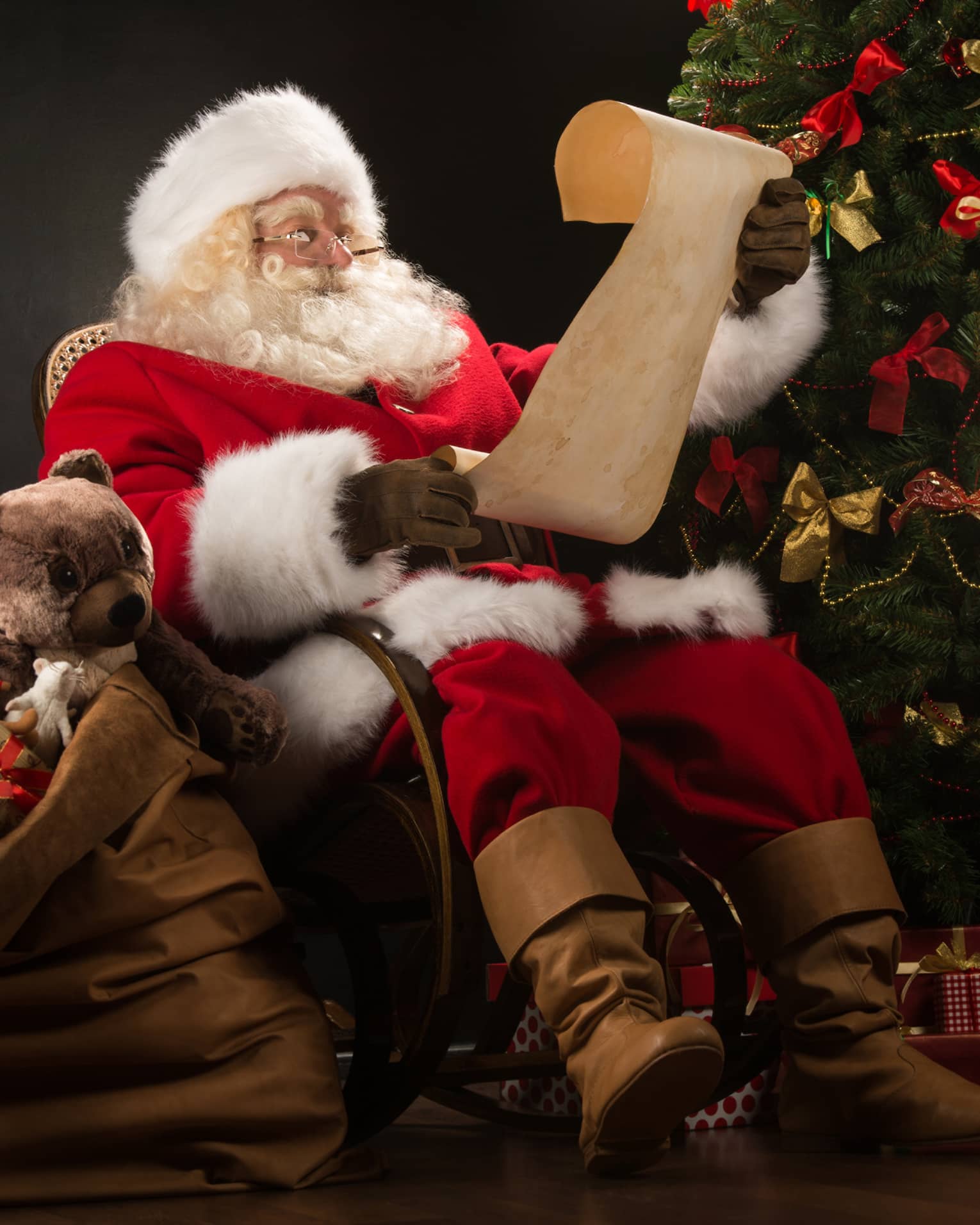 Santa Claus seated in large rocker holding a scroll, flanked by decorated Christmas tree with gifts and overflowing gift bag