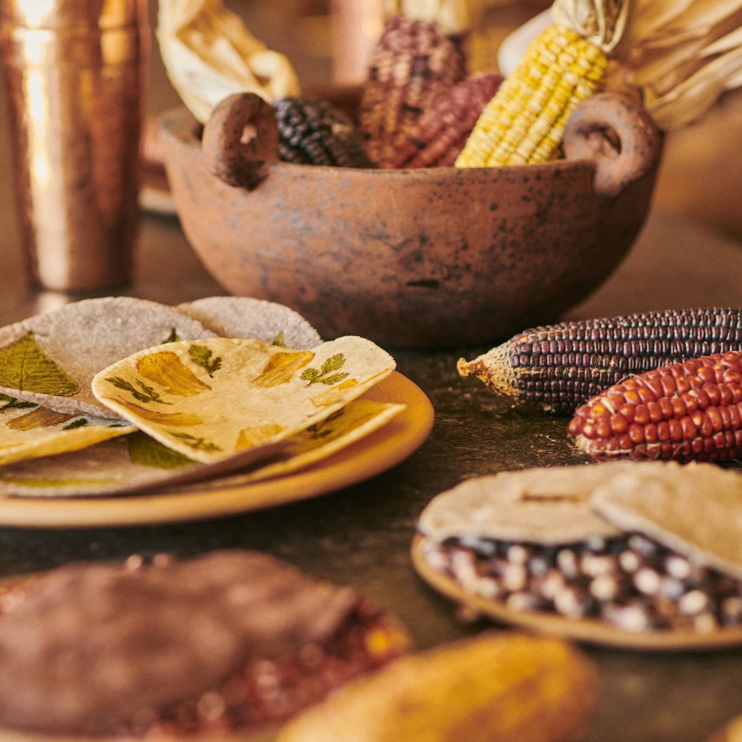 Close-up of copper cups, a rust-coloured stone bowl holding dried corn cobs and a yellow plate with hand-pressed tortillas.