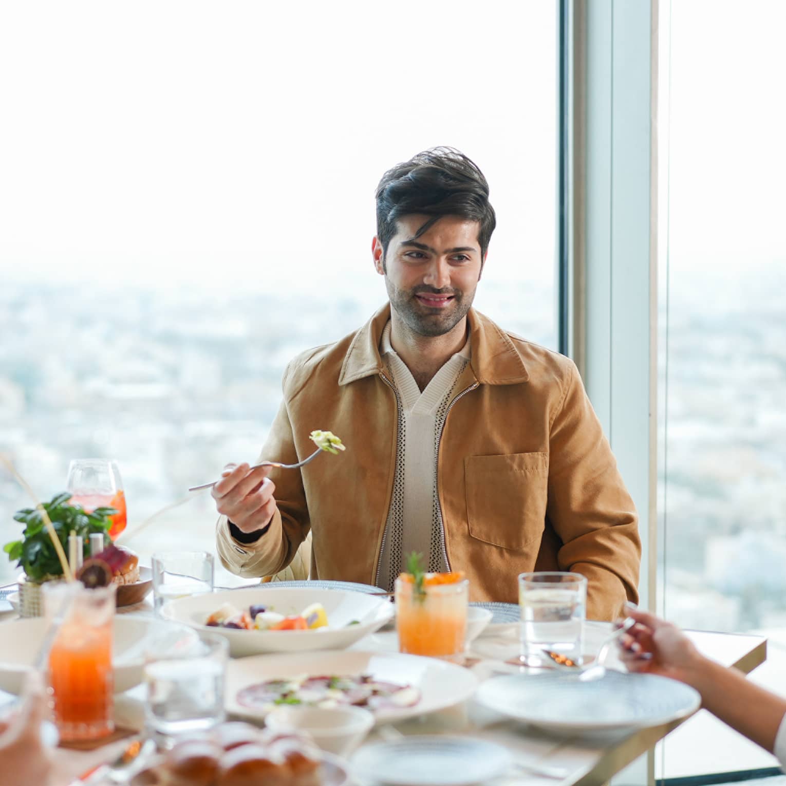 Man sits at restaurant table with a wall of windows behind him overlooking a city skyline