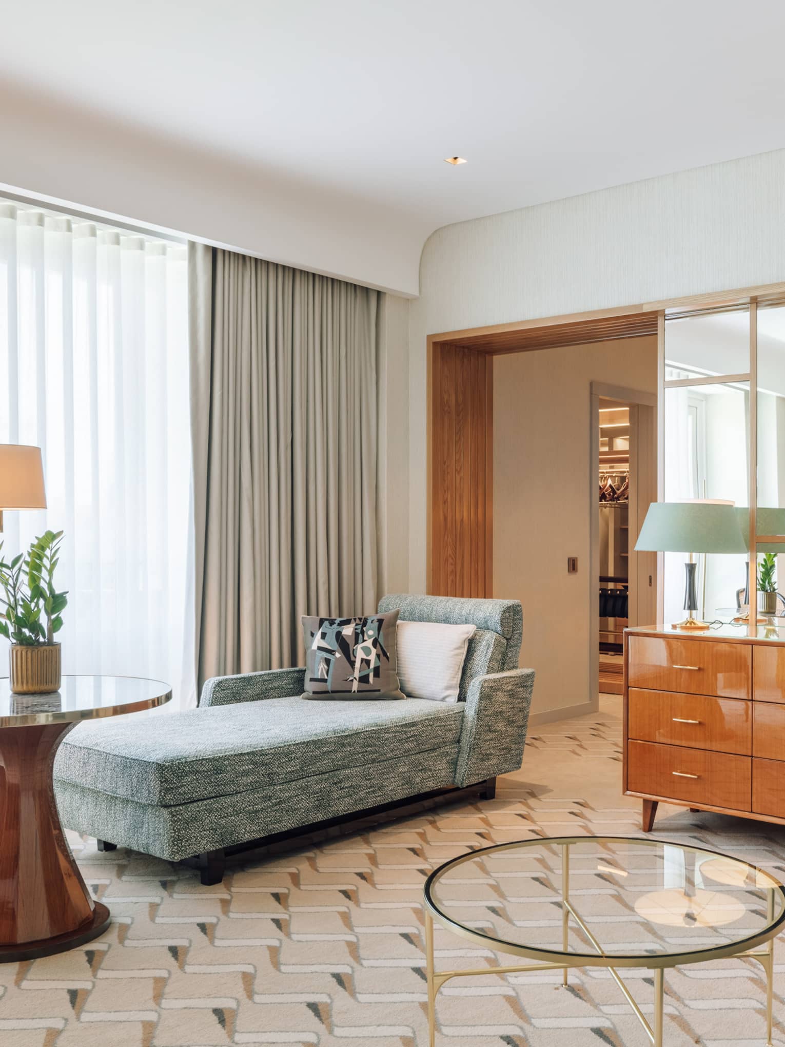 Hotel suite with a seating area, chaise lounge, round table and a bedroom partially divided by a dresser and shelving unit.