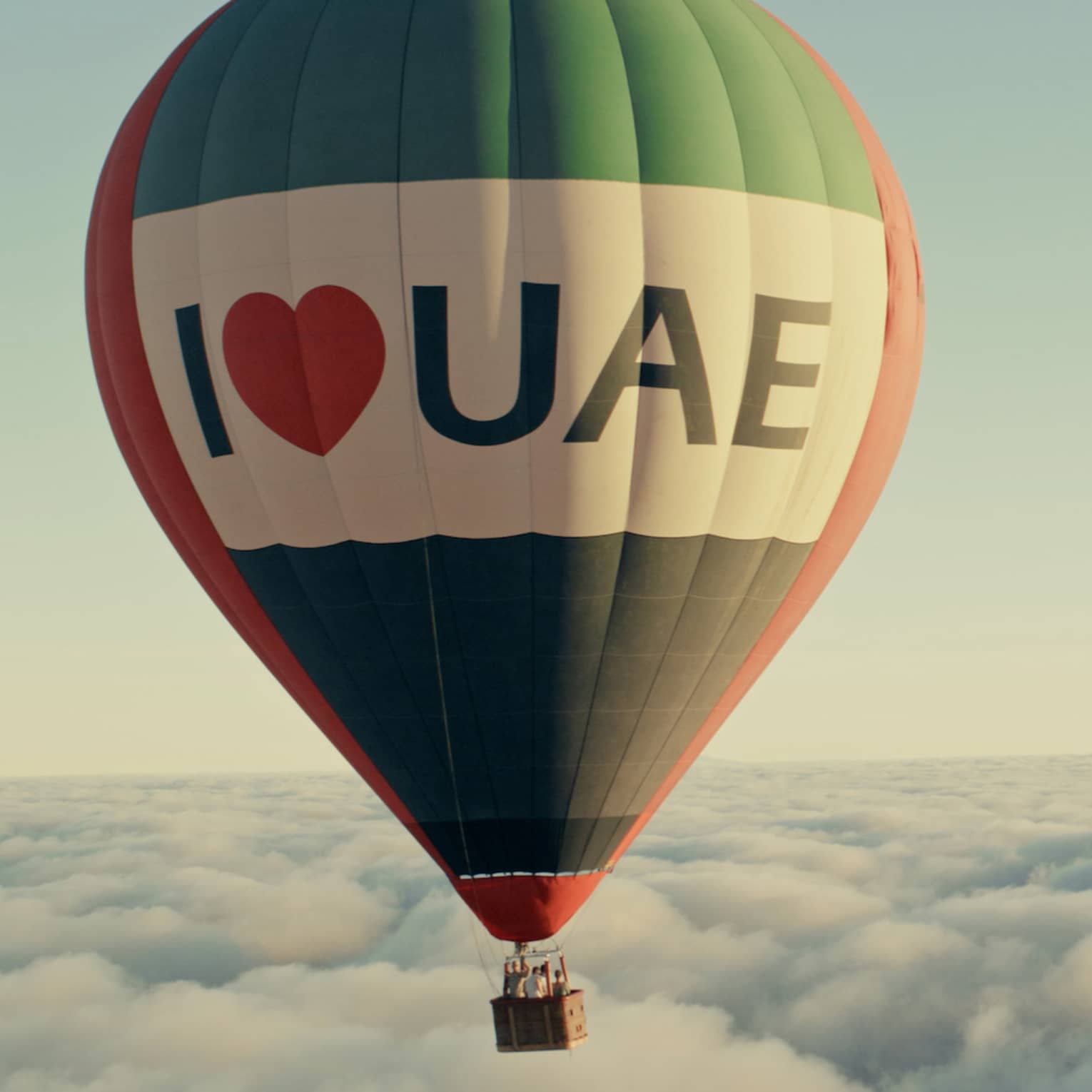 A hot air balloon flies above the clouds, in front of a light blue sky.
