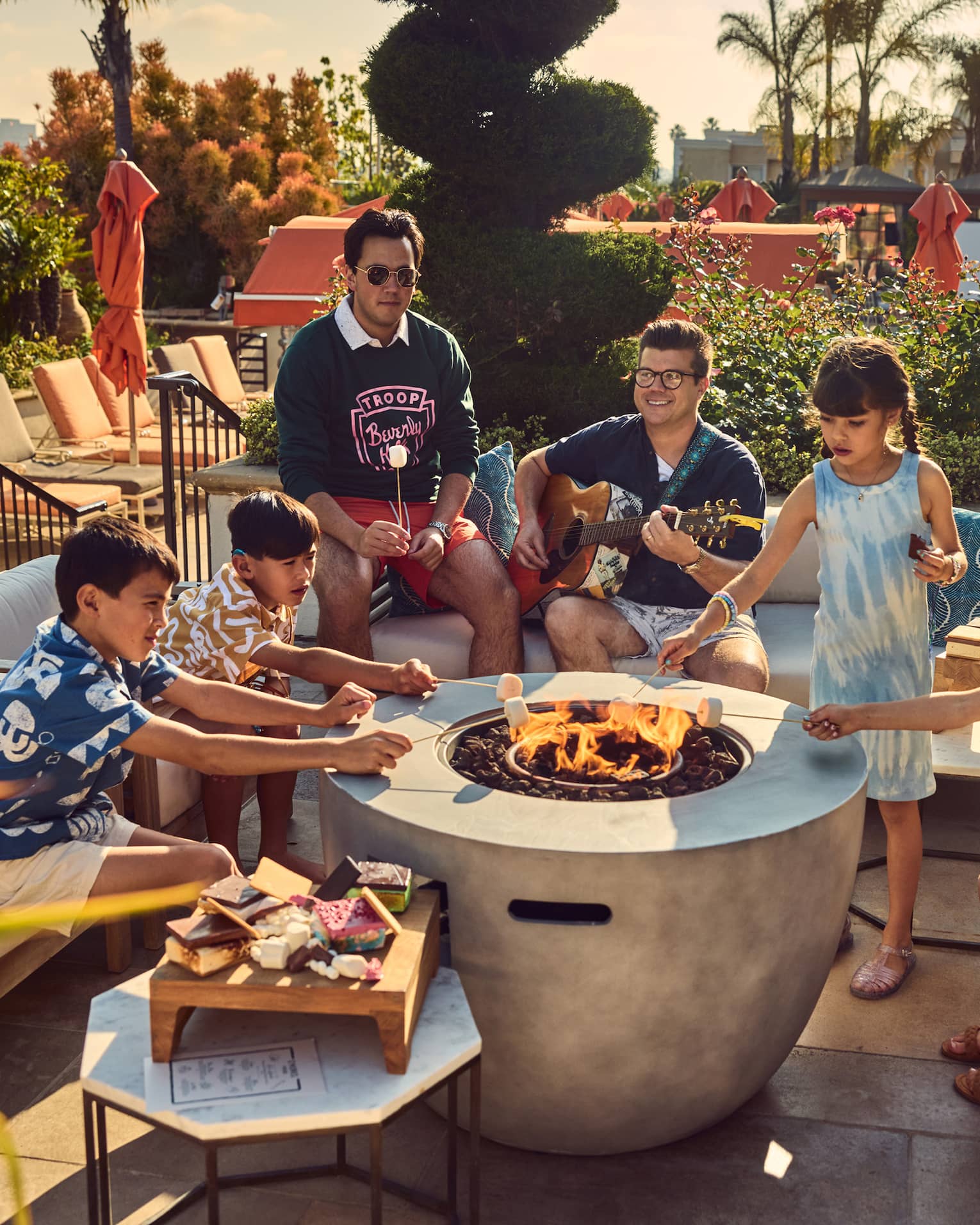 Young guests roasting marshmallows by a fire pit while a person plays the guitar