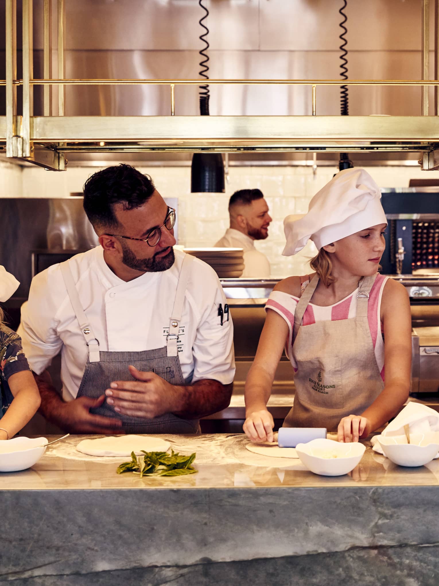 A chef standing at a counter teaching children wearing aprons and chef hats how to roll out pizza dough using rolling pins.