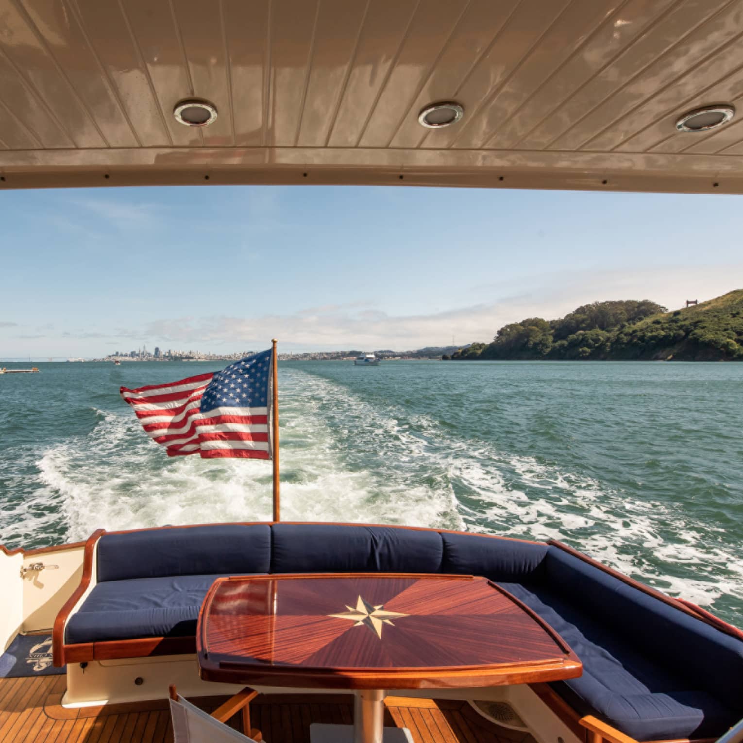 The back of a yacht, with a red table and an American flag, moving along the water.