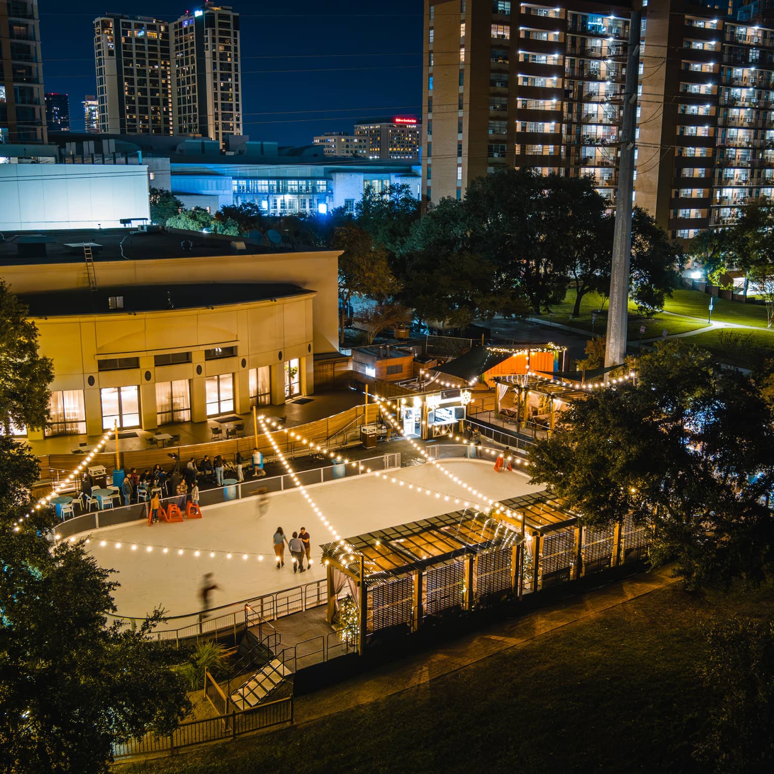 Aerial view of an outdoor ice rink with lights above and city buildings in the background.