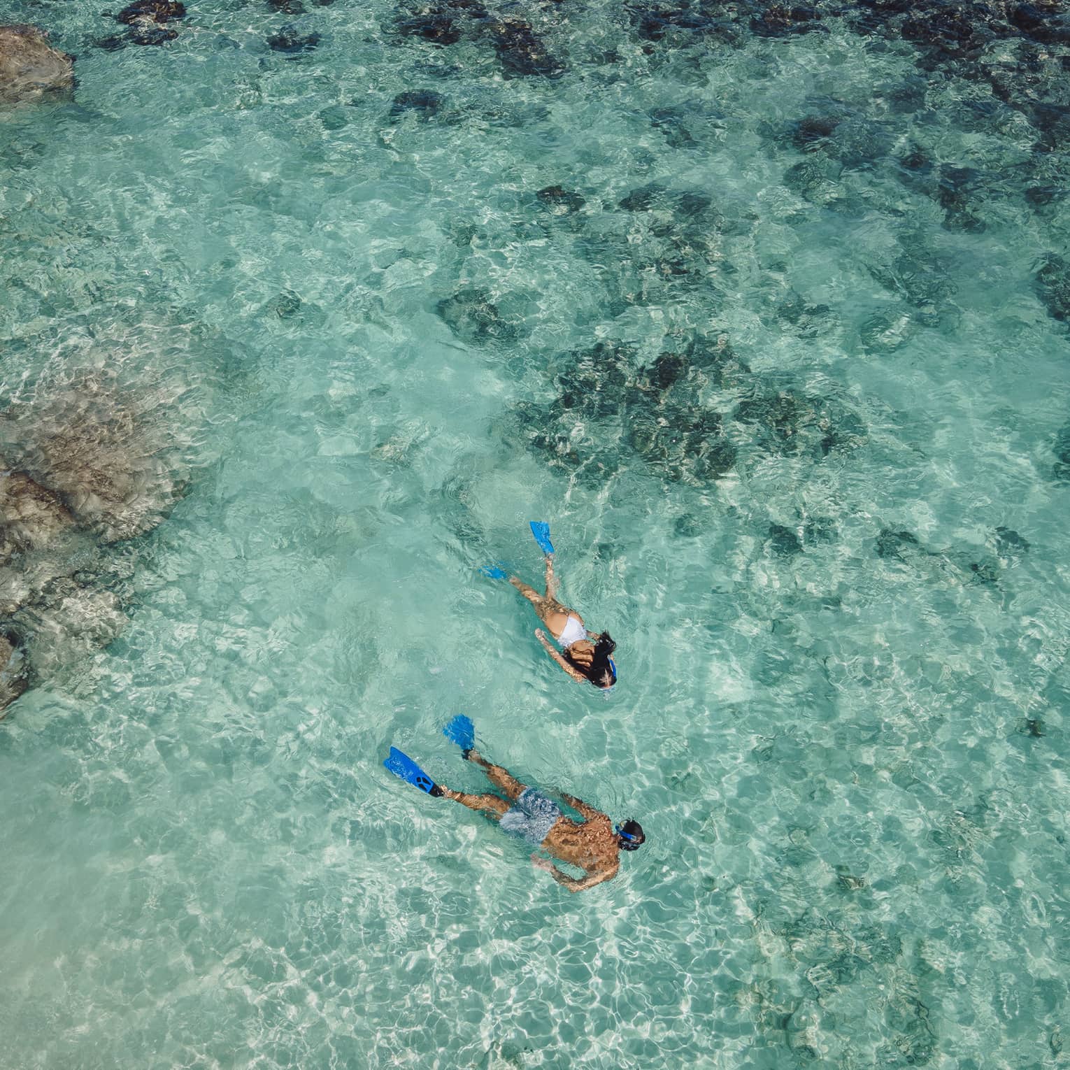 Aerial view of two snorkellers wearing blue flippers in an expanse of shallow, turquoise waters rippling over the reef.