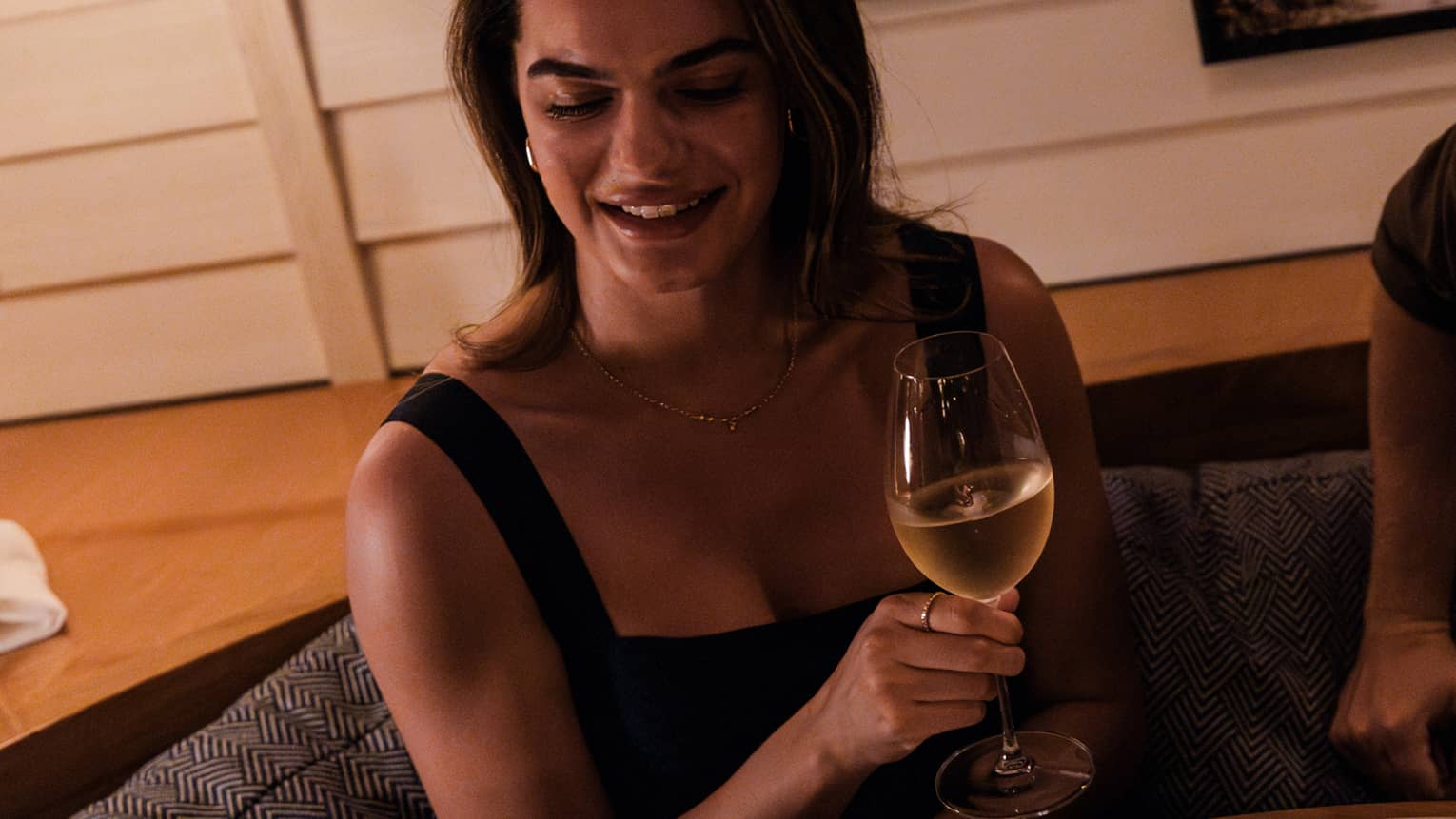 Guests sit at a booth table in a dining space. A person is handing the guests a plate of food, while the guest holds a glass of wine in a stemmed glass.