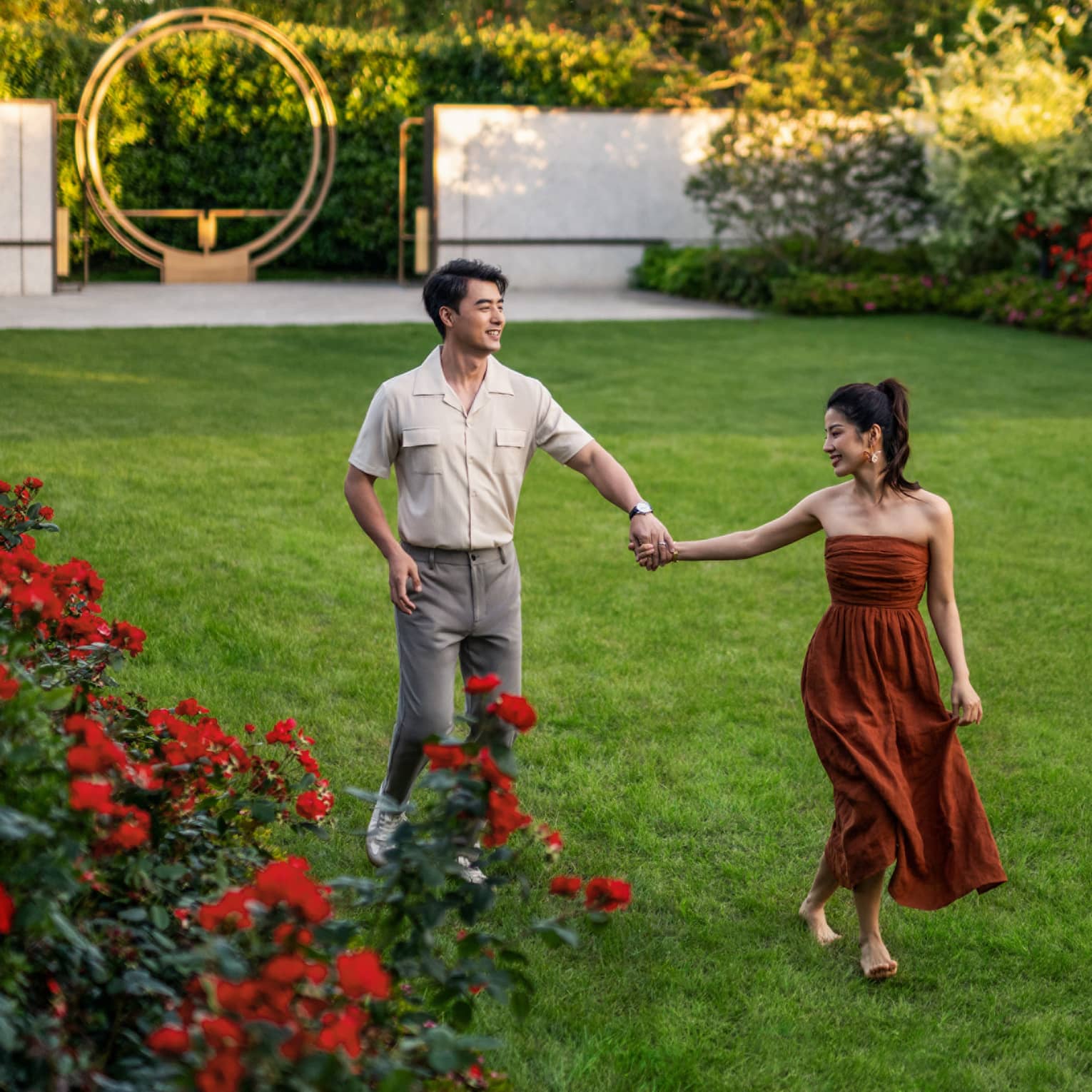 A couple holding hands crosses a thick lawn bordered by trees and bright red and fucsia blooms, and a metal abstract artwork.