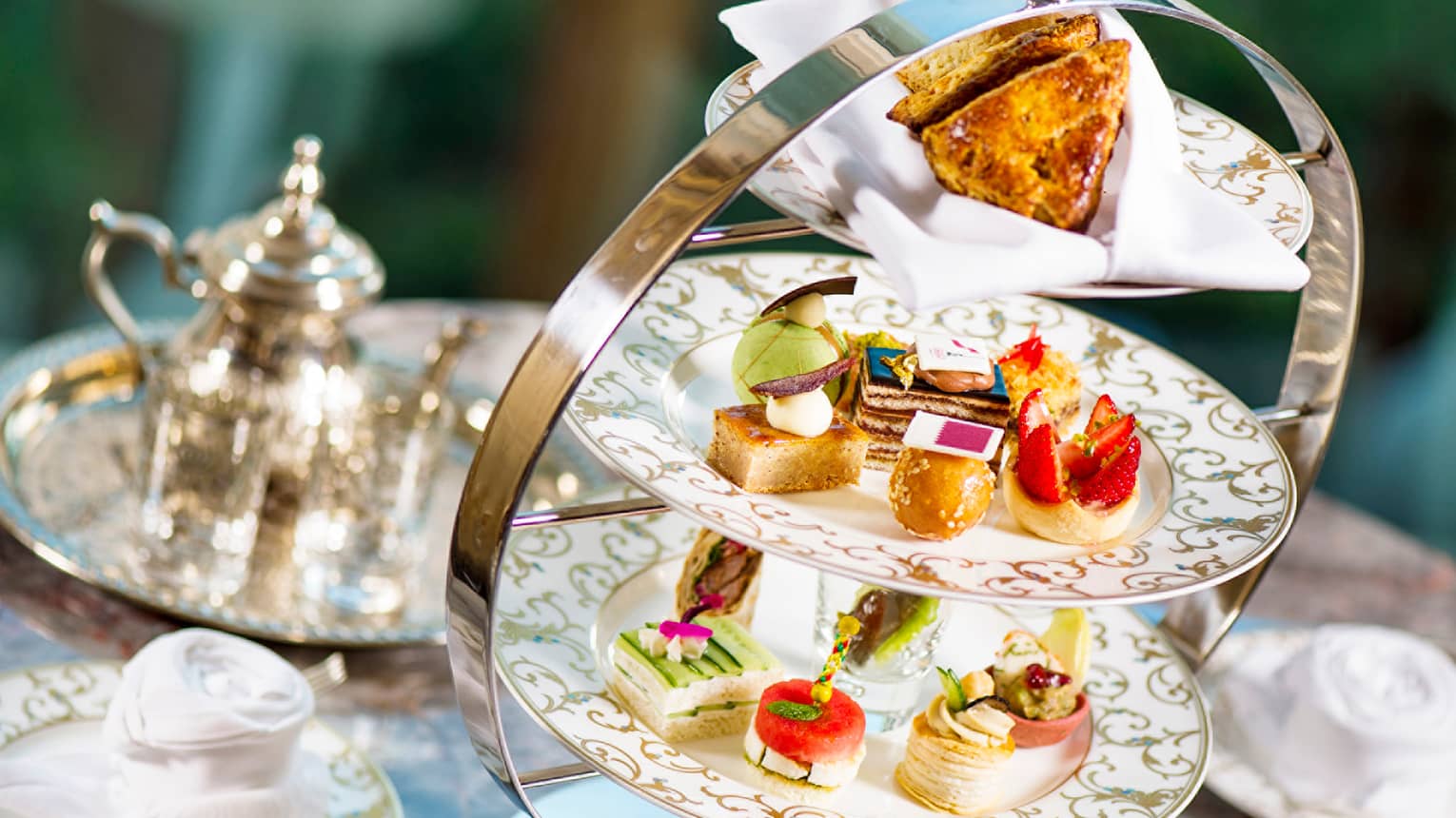 Small, artful pastries displayed on gold-accented china plates in a three-tier stand. Behind, a silver teapot and tray.