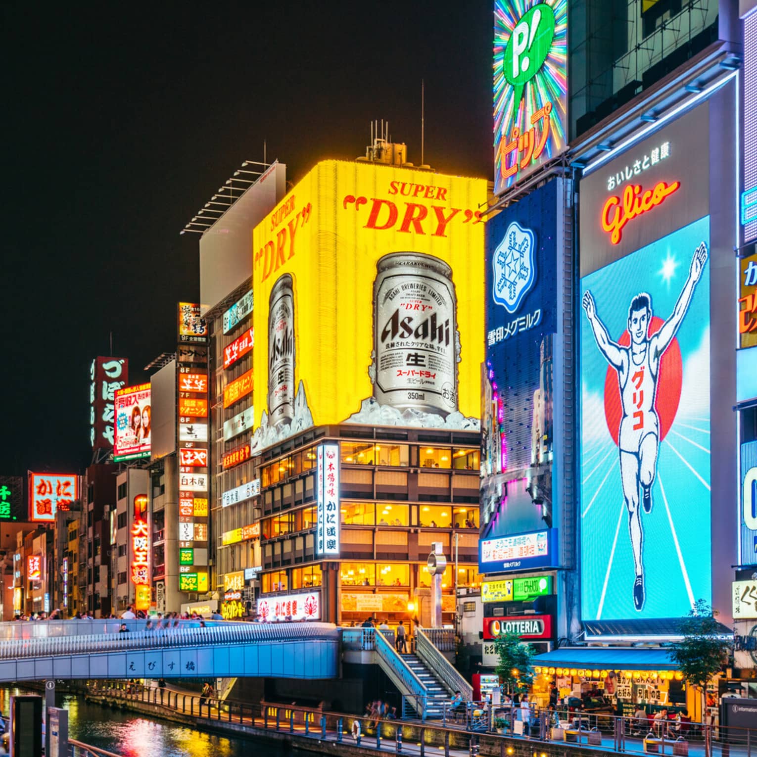 A footbridge crosses a narrow canal flanked by huge neon ads in bright colours on tall buildings in a busy city at night.